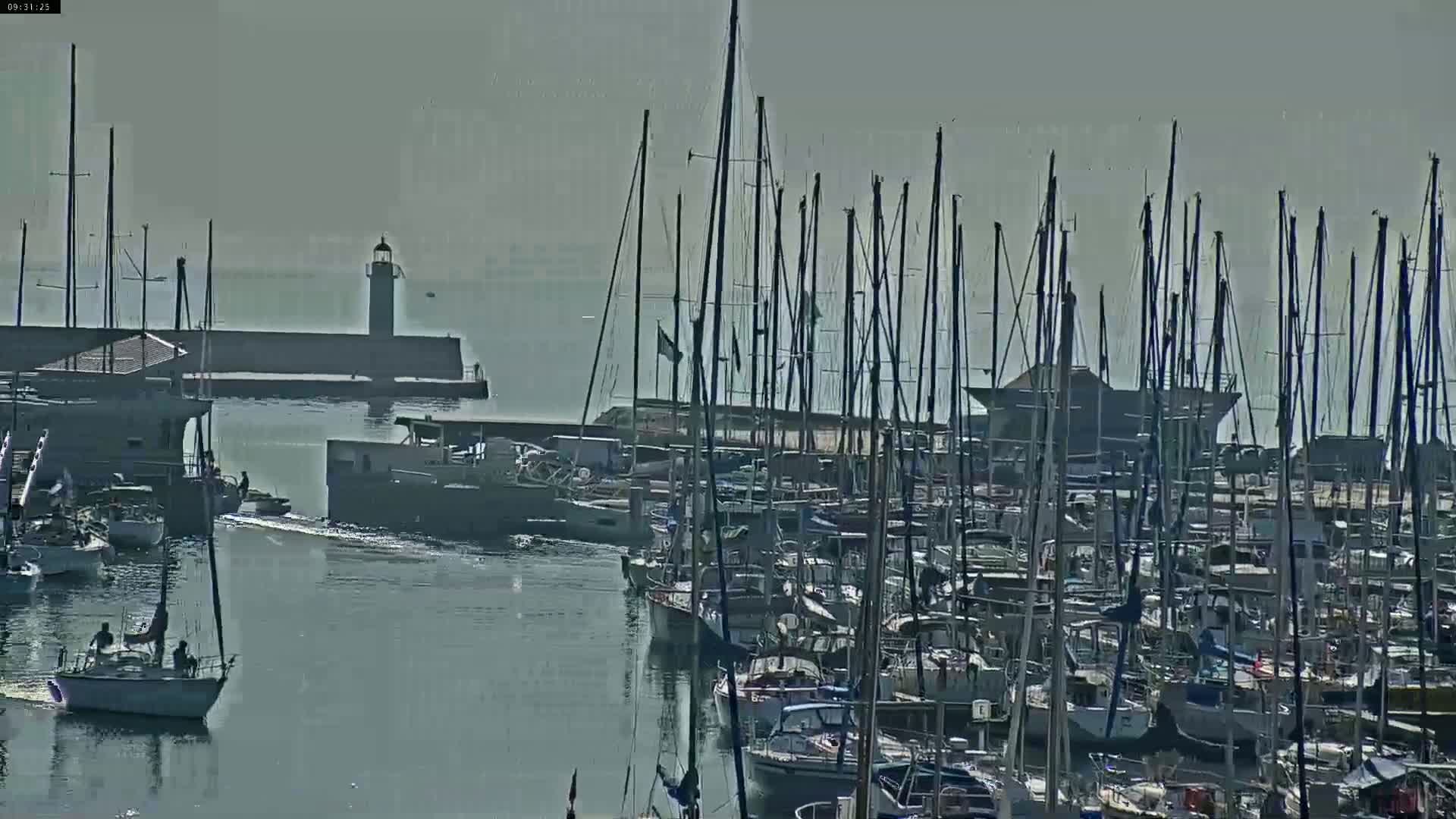 A harbor filled with numerous sailboats under a hazy sky, with a lighthouse visible in the distance.