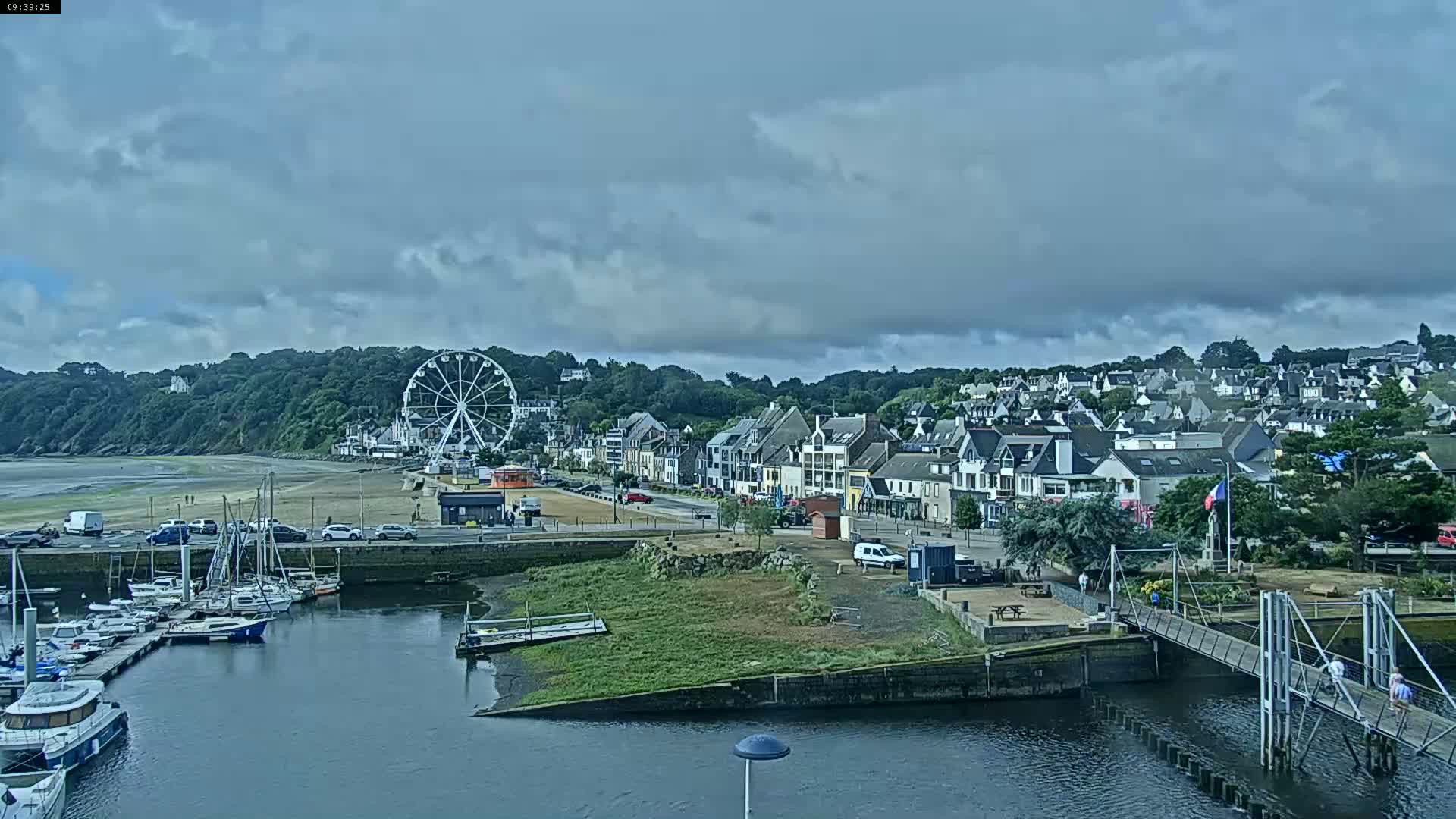 A coastal town with a Ferris wheel is visible under a cloudy sky, showcasing a harbor filled with boats and a pedestrian bridge leading to a grassy area.