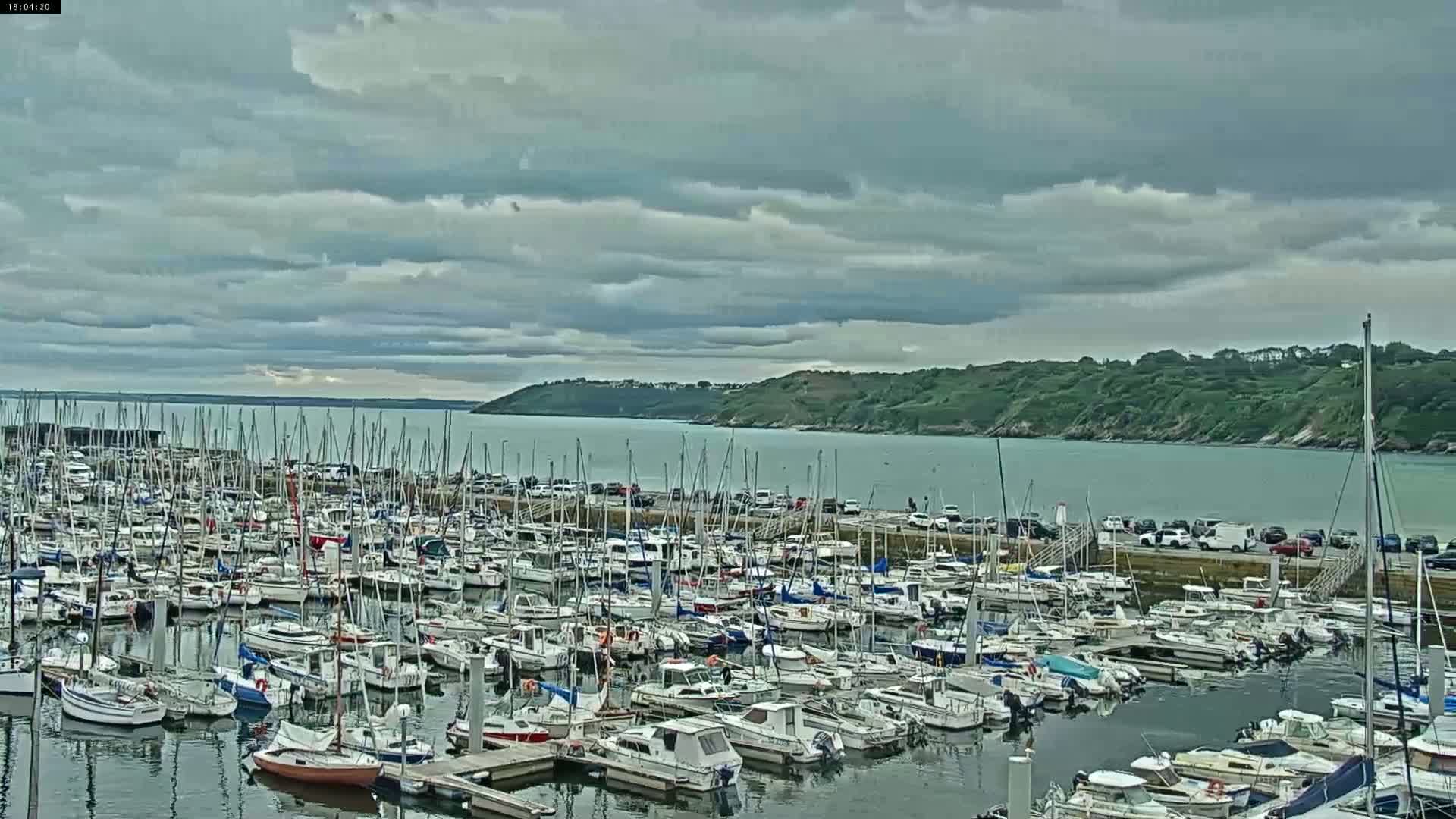 A coastal town with a Ferris wheel is visible under a cloudy sky, showcasing a harbor filled with boats and a pedestrian bridge leading to a grassy area.