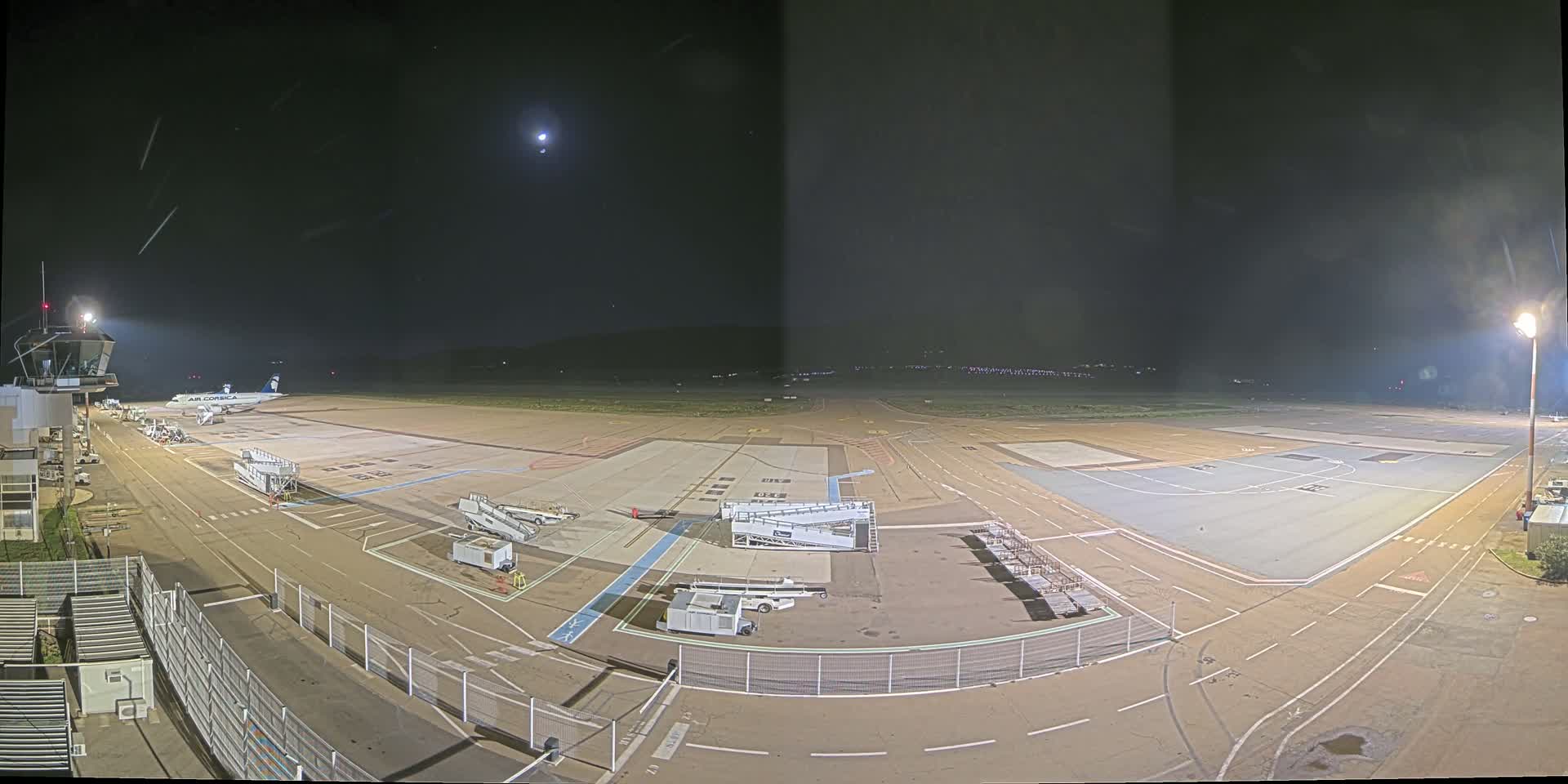 A nighttime view of an airport tarmac with a single airplane parked at a gate under a mostly clear sky with a visible moon.