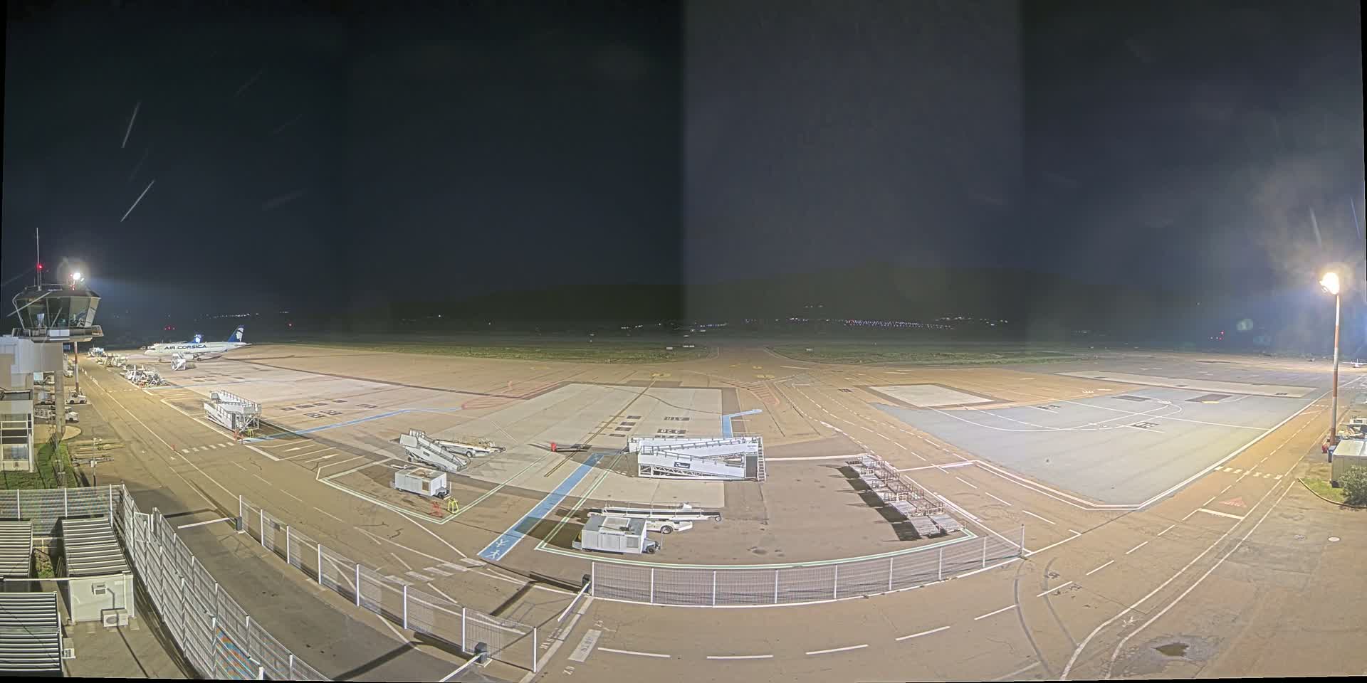 A nighttime view of an airport tarmac with a single airplane parked near a gate, various ground support equipment, and an air traffic control tower visible.
