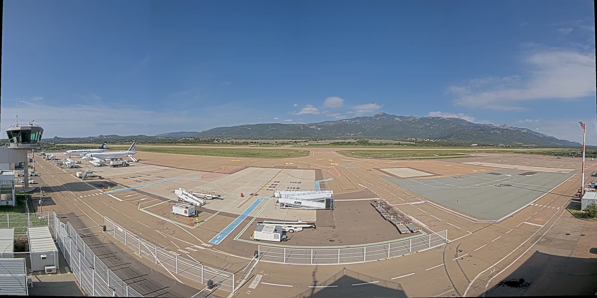 A sunny day at an airport with two airplanes parked on the tarmac, surrounded by ground support equipment, with mountains in the background.
