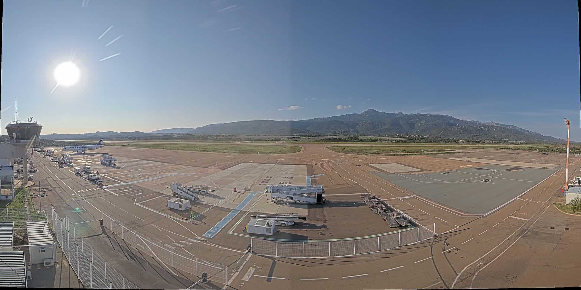 A sunny day at an airport with a plane parked at the gate,  ground support equipment, and mountains in the background.