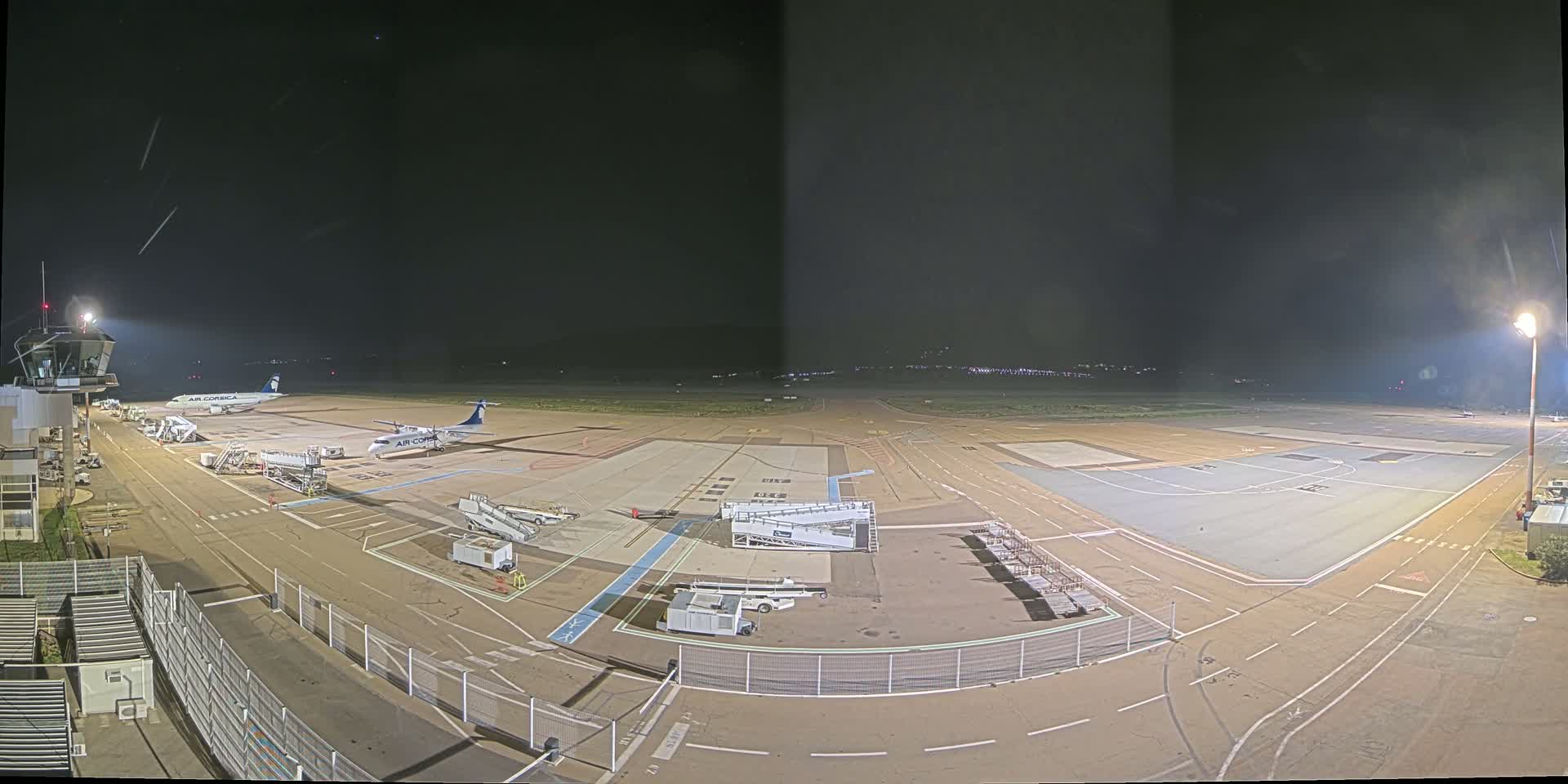 A nighttime view of an airport tarmac with two airplanes parked near the terminal, surrounded by service vehicles and under bright lights.