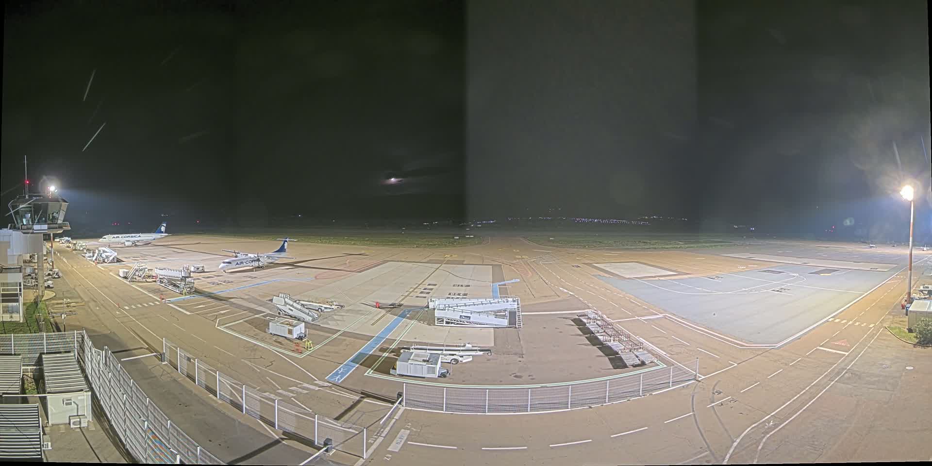 A nighttime view of an airport tarmac with two airplanes parked at gates, surrounded by ground support equipment and brightly lit.