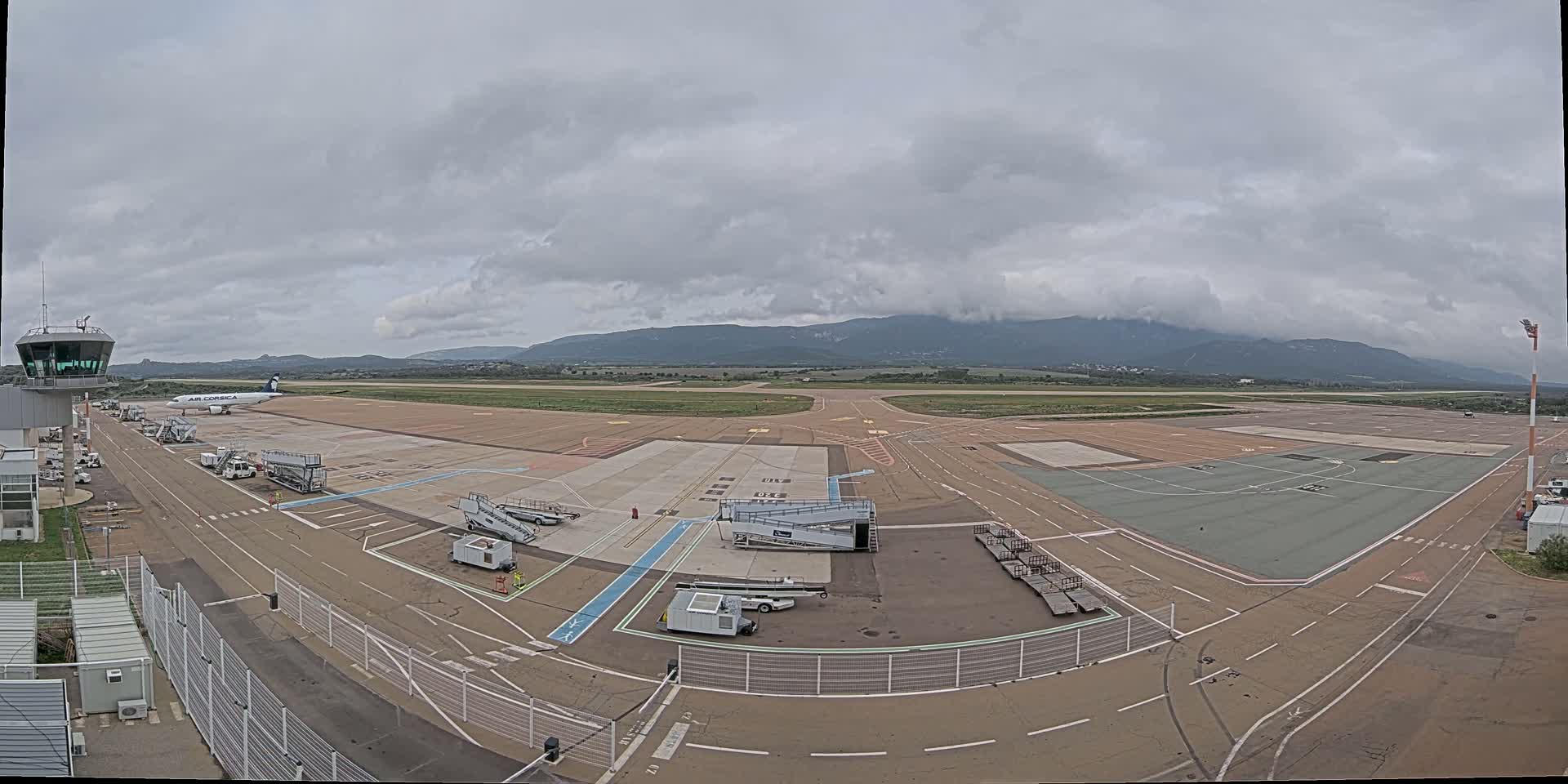 An airport tarmac on an overcast day, with a single airplane and several ground support vehicles visible, mountains in the distance.