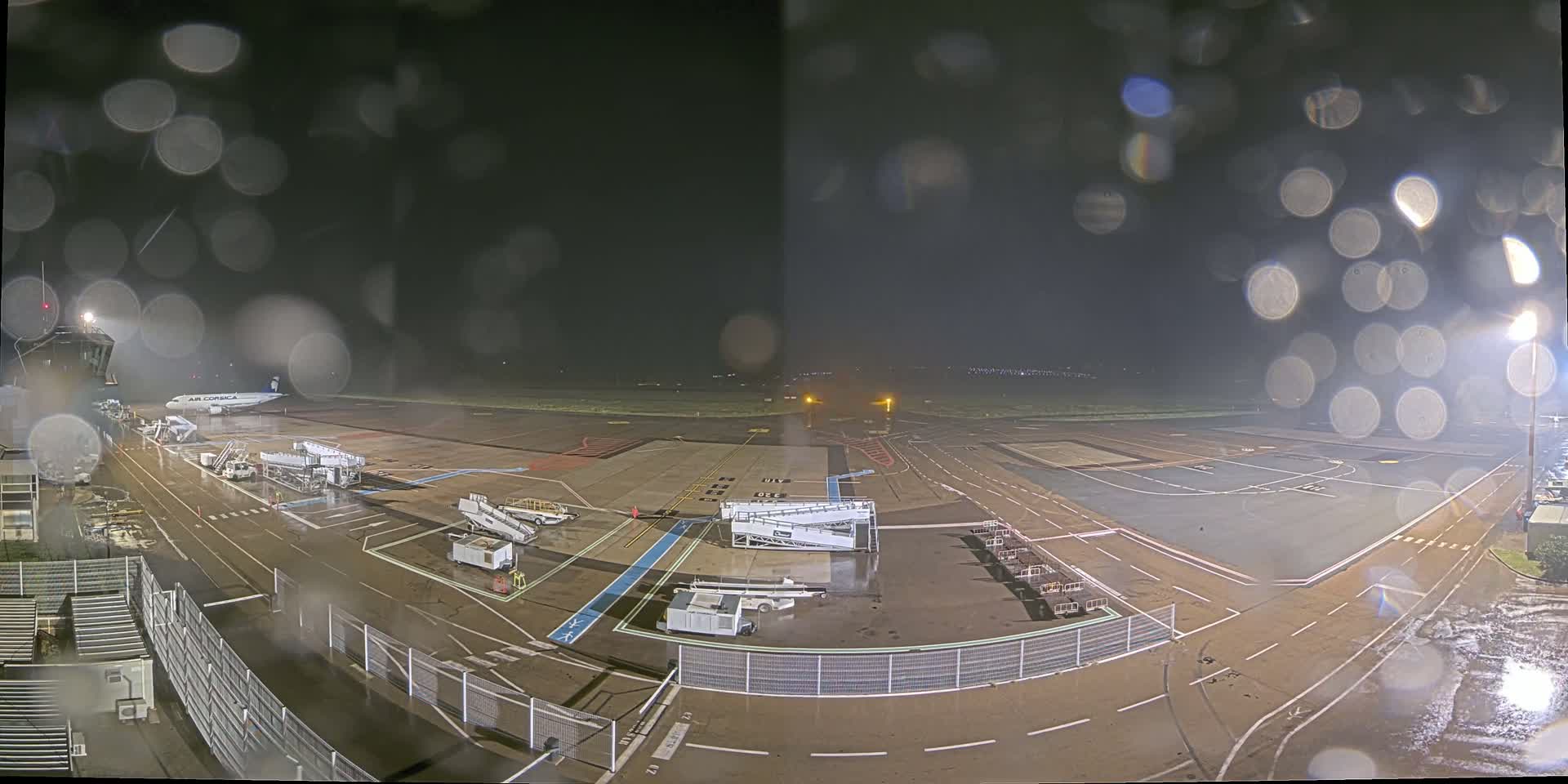 A nighttime view of an airport tarmac in the rain, showing an airplane at a gate with ground support equipment.