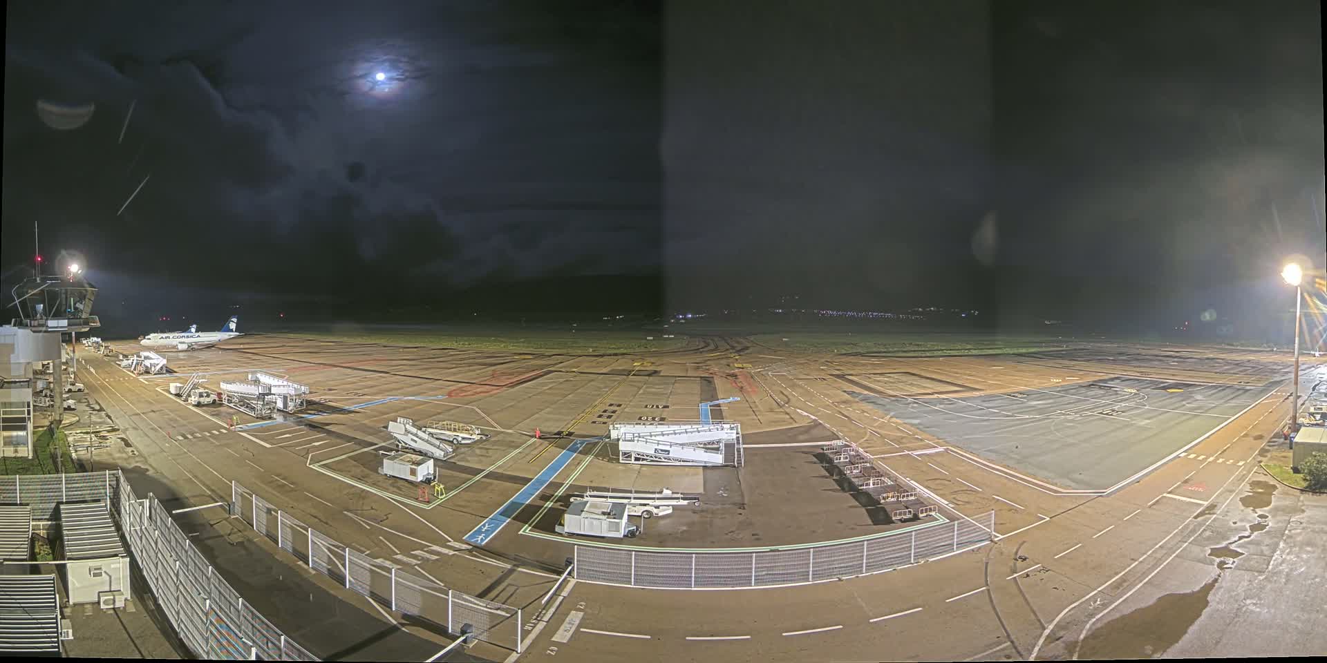A nighttime view of an airport tarmac under a partly cloudy moonlit sky, showing a single airplane parked near a terminal building with ground support equipment and jet bridges.
