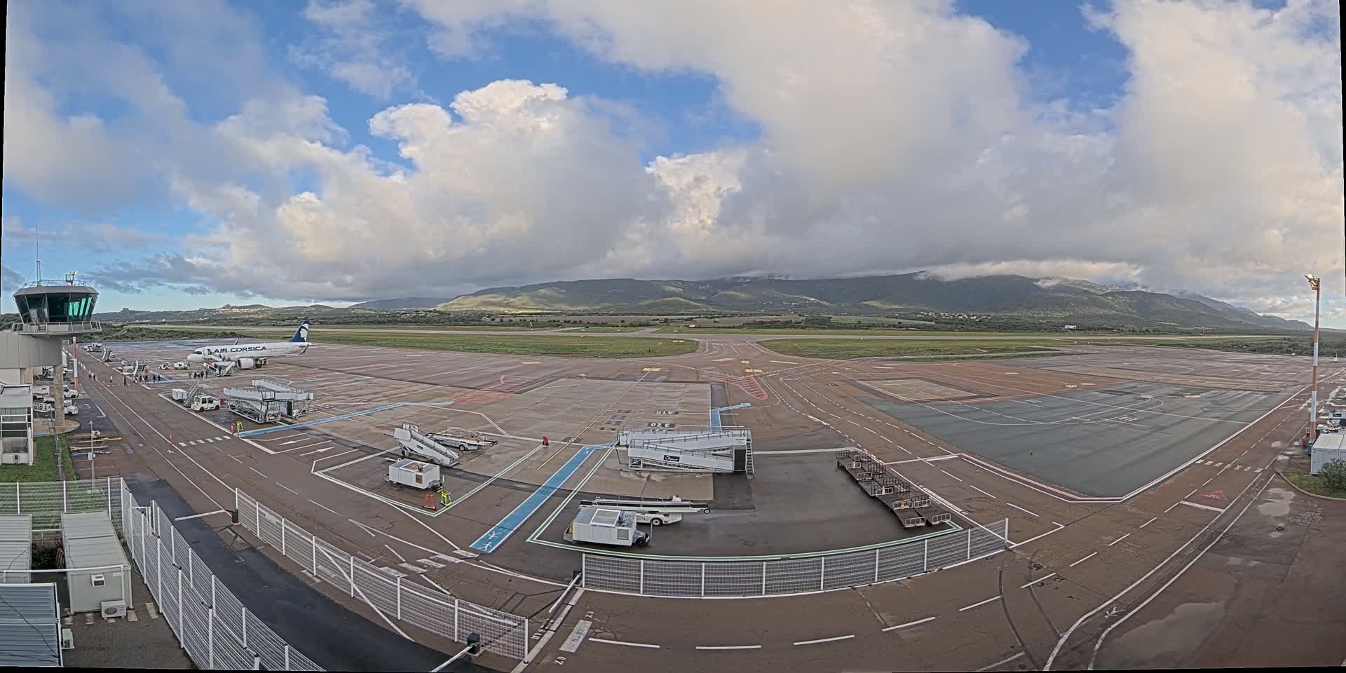 An airport tarmac with a plane at the gate, ground support equipment, and a control tower, under a partly cloudy sky with mountains in the background.