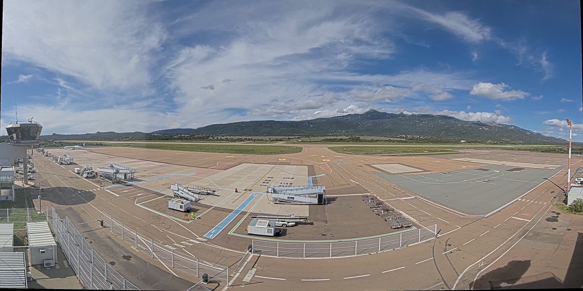 A mostly sunny day at an airport with several jet bridges and aircraft parked on the tarmac, backed by a mountain range in the distance.