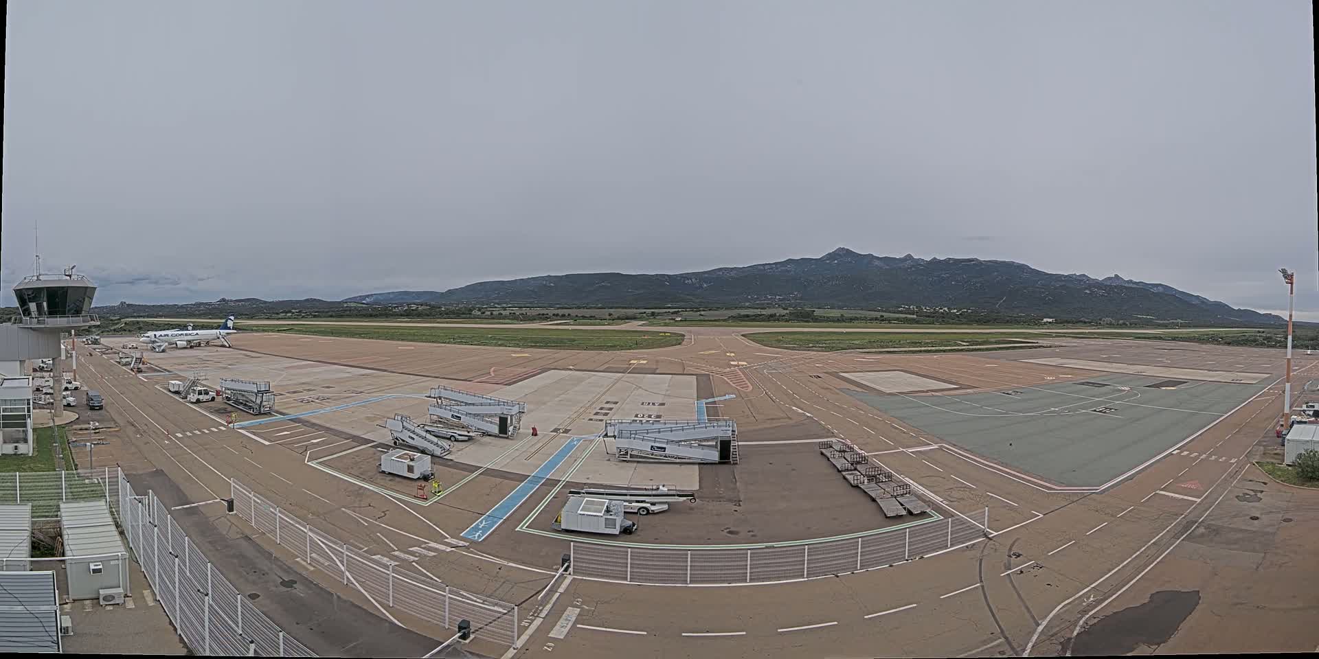 An airport tarmac with a single airplane and several ground support vehicles is visible under an overcast sky, with mountains in the background.