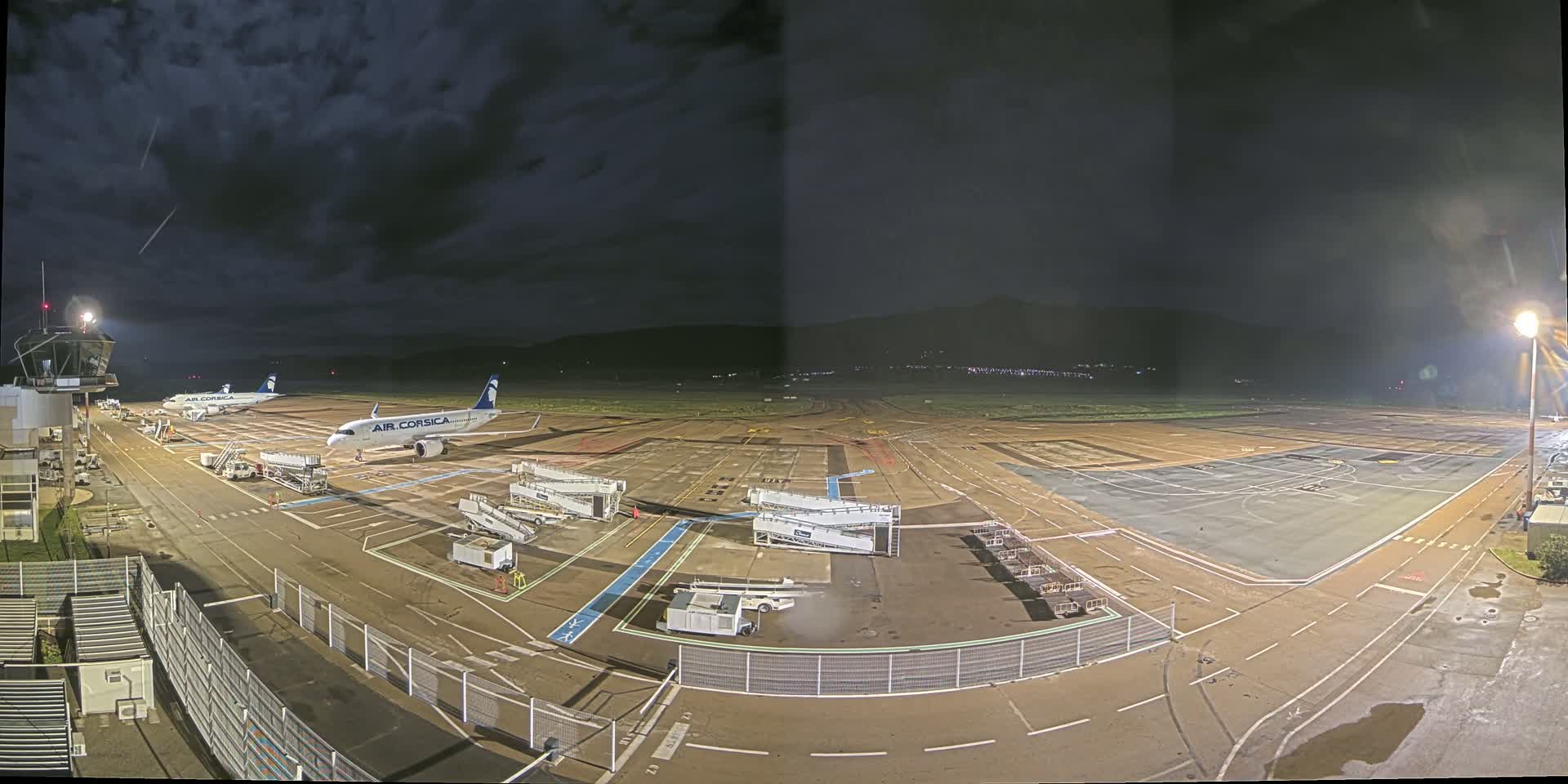 A nighttime view of an airport tarmac with two airplanes parked near the terminal building under a dark, cloudy sky.