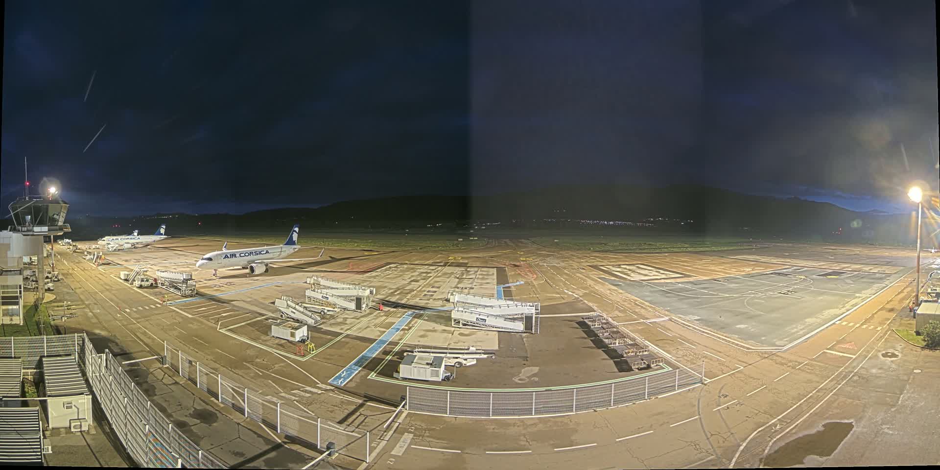 At night, an airport tarmac shows two airplanes parked at gates, surrounded by ground support equipment, under a dark sky.