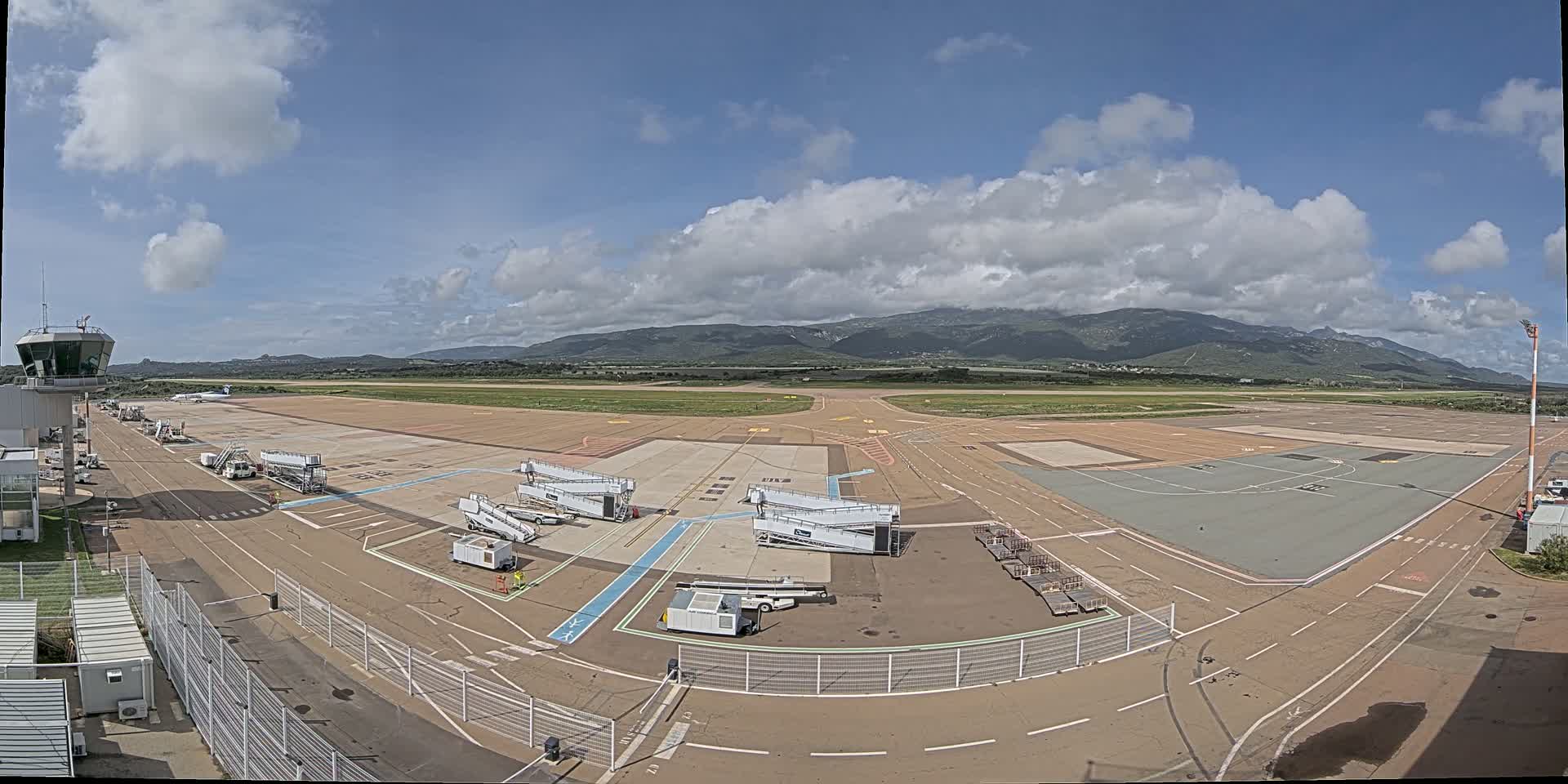 An airport tarmac with several jet bridges and ground support equipment, under a partly cloudy sky with mountains visible in the distance.