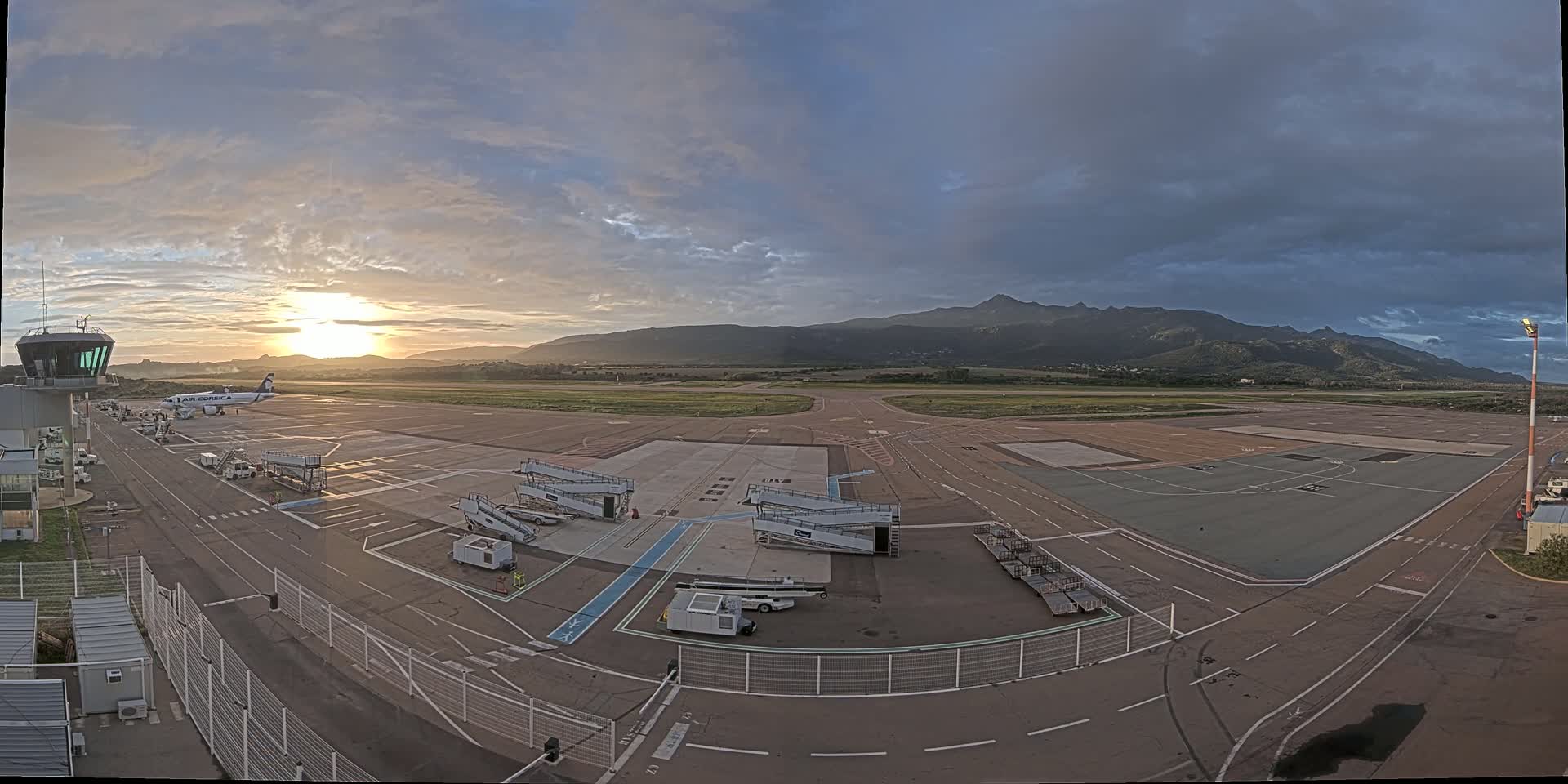 An airport tarmac at sunrise with a single airplane parked at a gate, under partly cloudy skies and with mountains in the background.