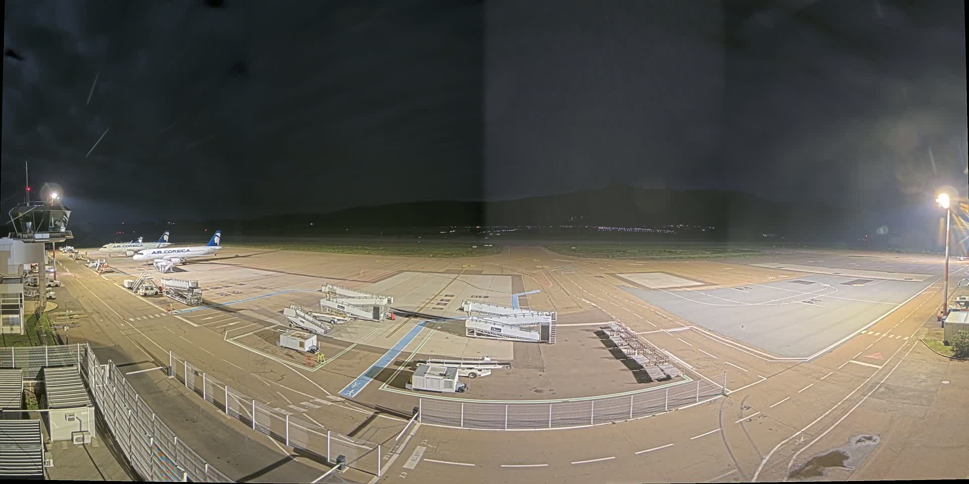A nighttime view of an airport tarmac with two airplanes parked at gates, ground support equipment, and a control tower, under a dark sky.