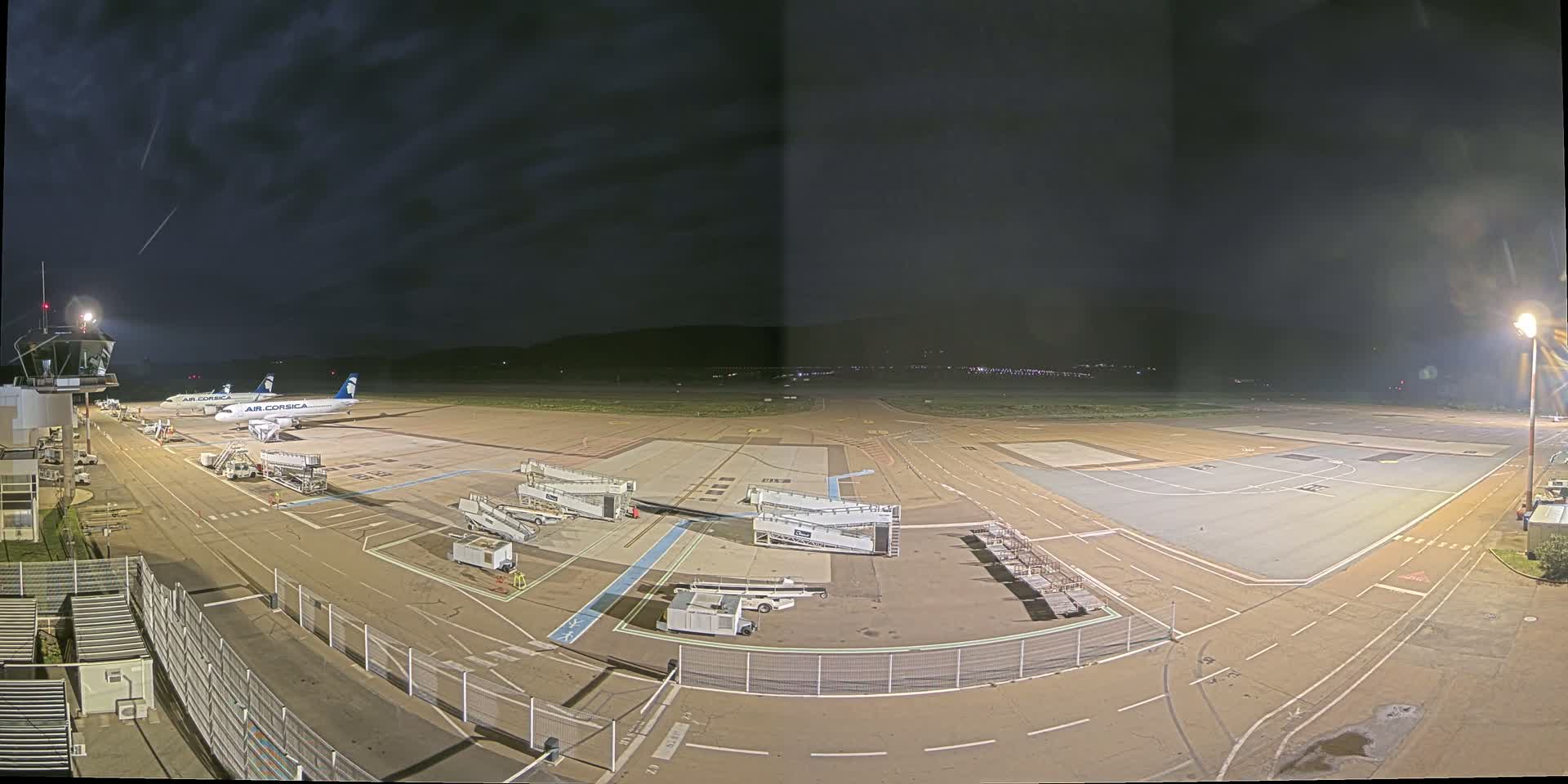 A nighttime view of an airport tarmac with two airplanes parked at gates, surrounded by ground support equipment under a dark, cloudy sky.