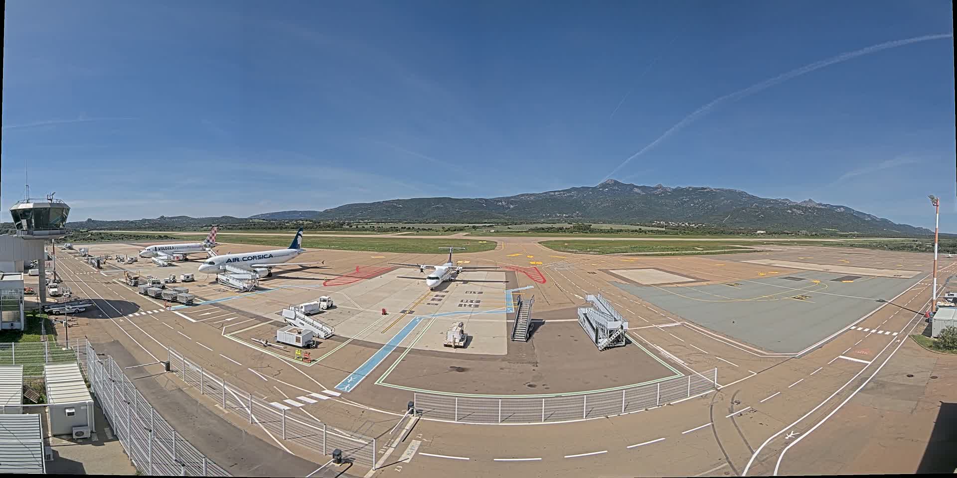 An airport tarmac under a clear blue sky with mountains in the distance, showing several airplanes, ground support equipment, and an air traffic control tower.
