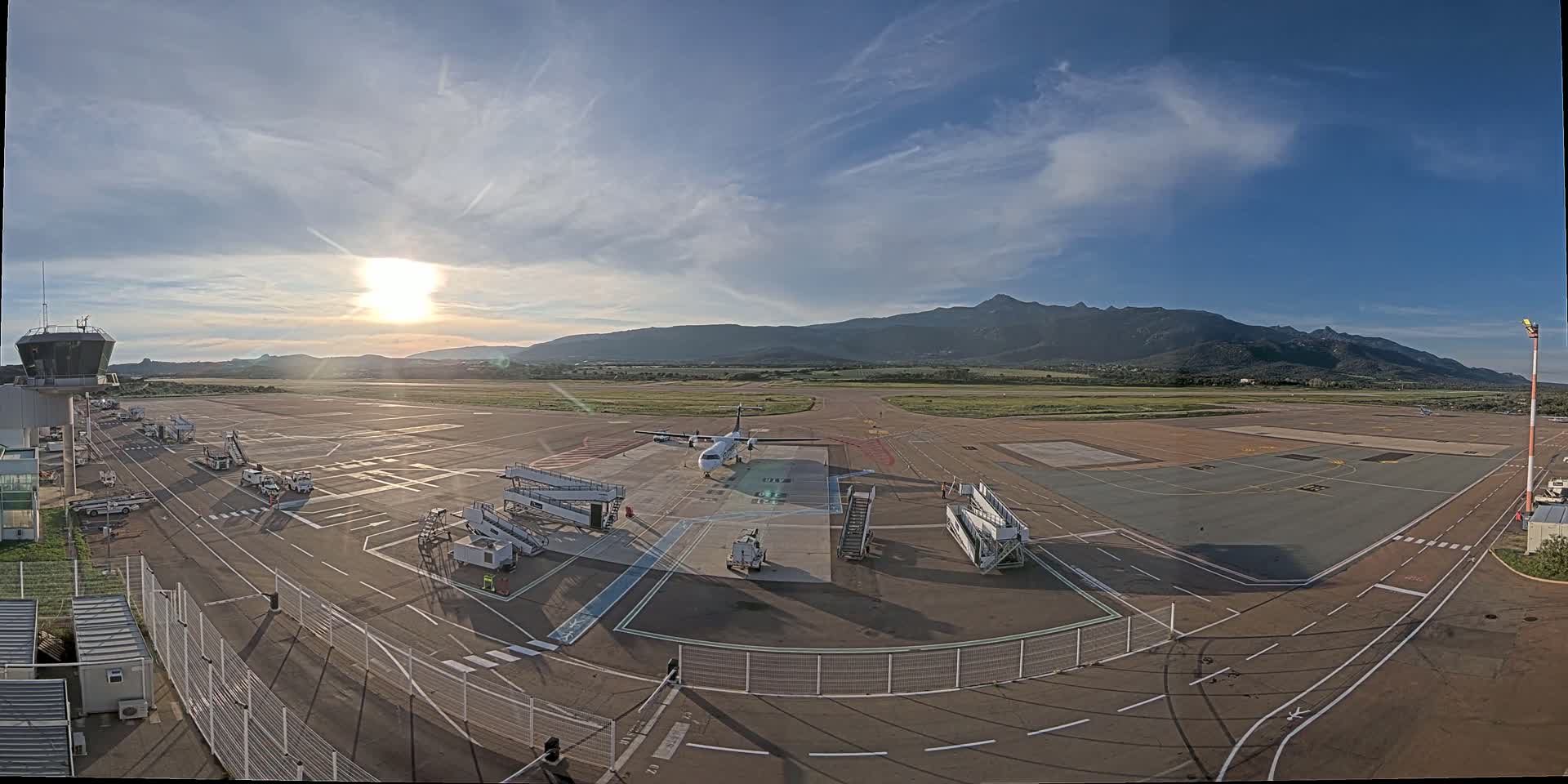 A mostly sunny day at an airport shows a plane at a gate with jet bridges and ground support equipment, all in front of a mountain range.