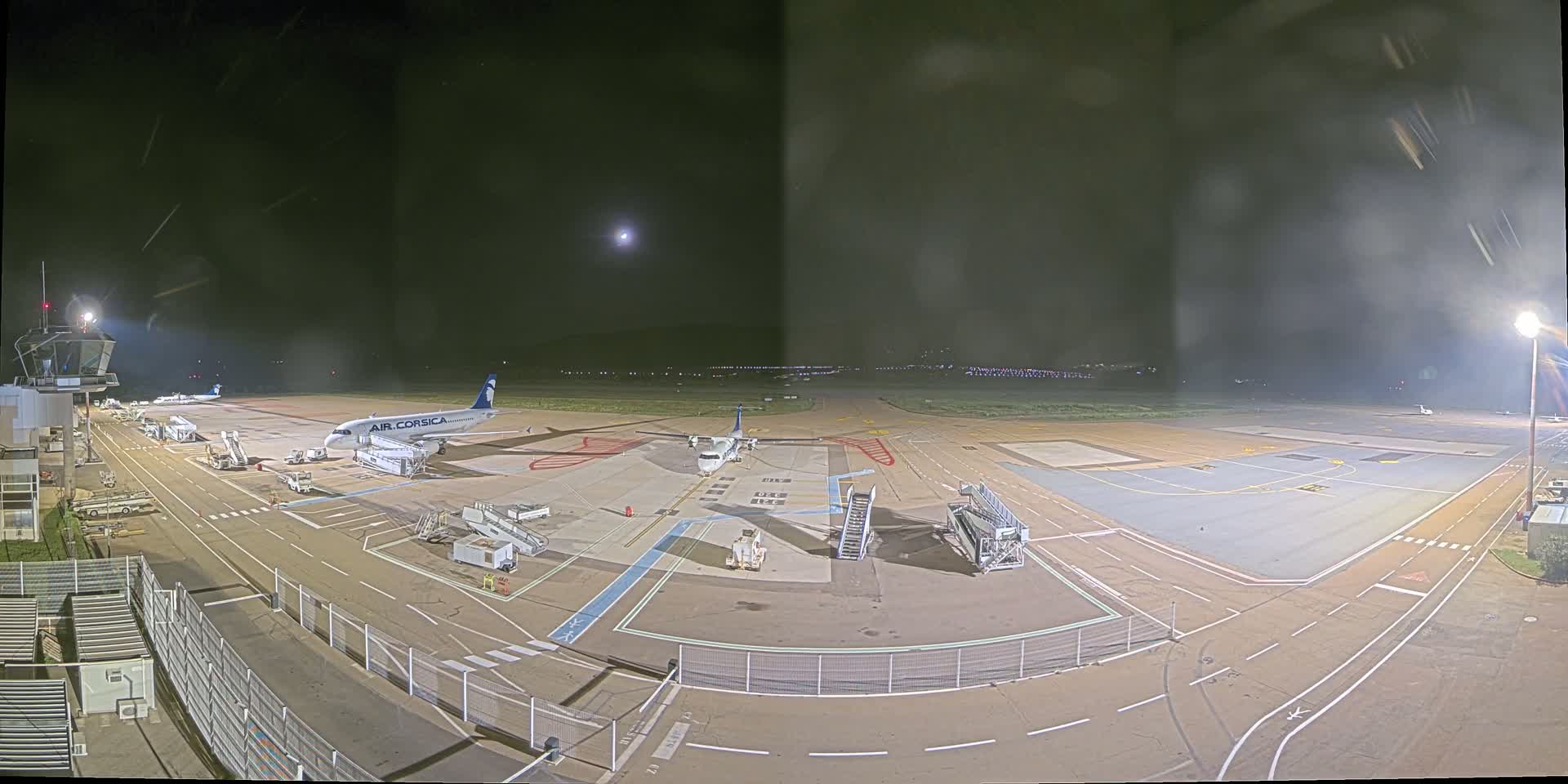 At night, a mostly empty airport tarmac shows two airplanes parked at gates with ground support equipment, and a control tower visible in the distance under a mostly clear sky with a bright moon.