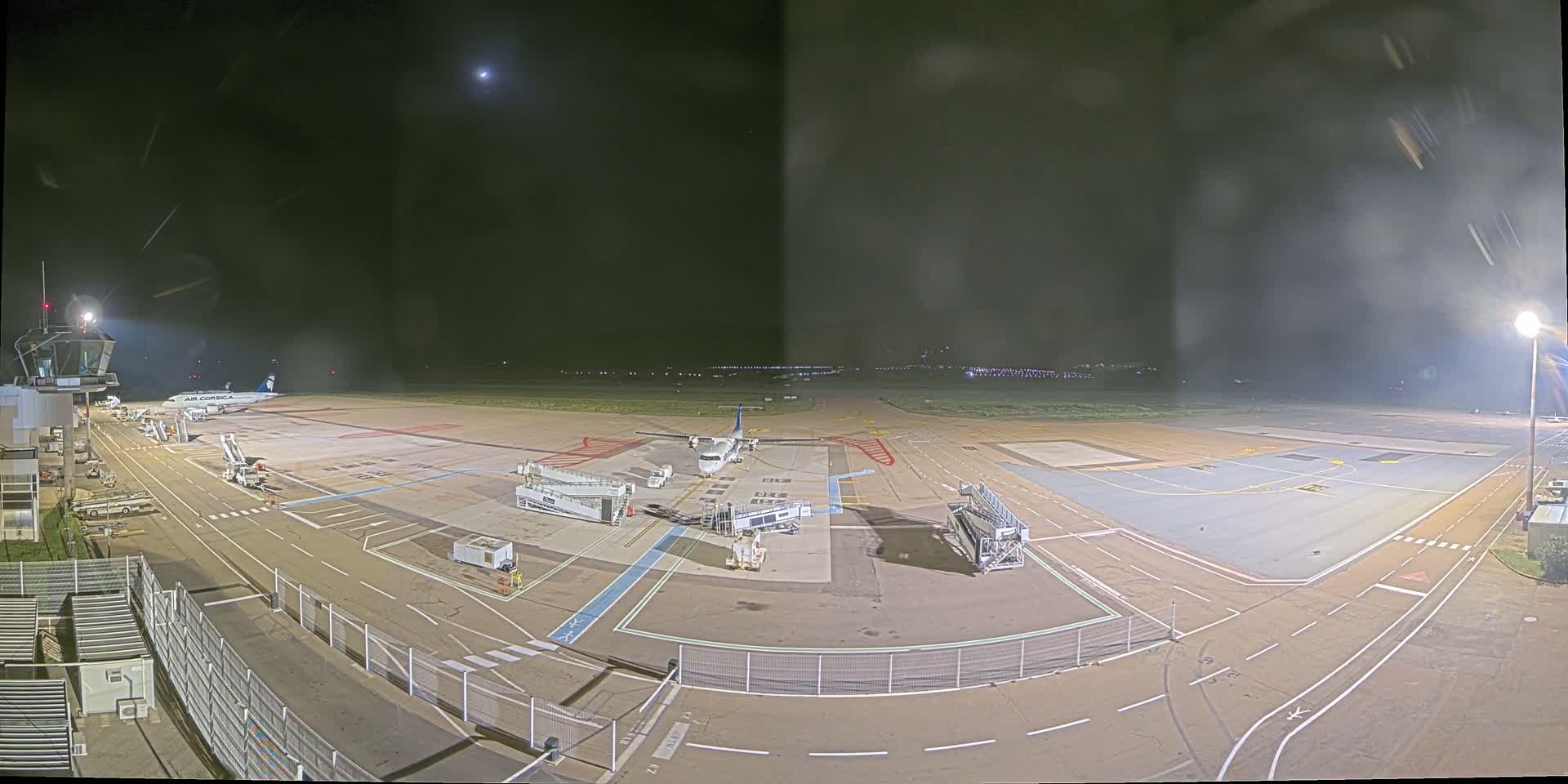 A nighttime view of an airport tarmac with two airplanes parked at gates, surrounded by ground support equipment and brightly lit under a mostly clear sky.