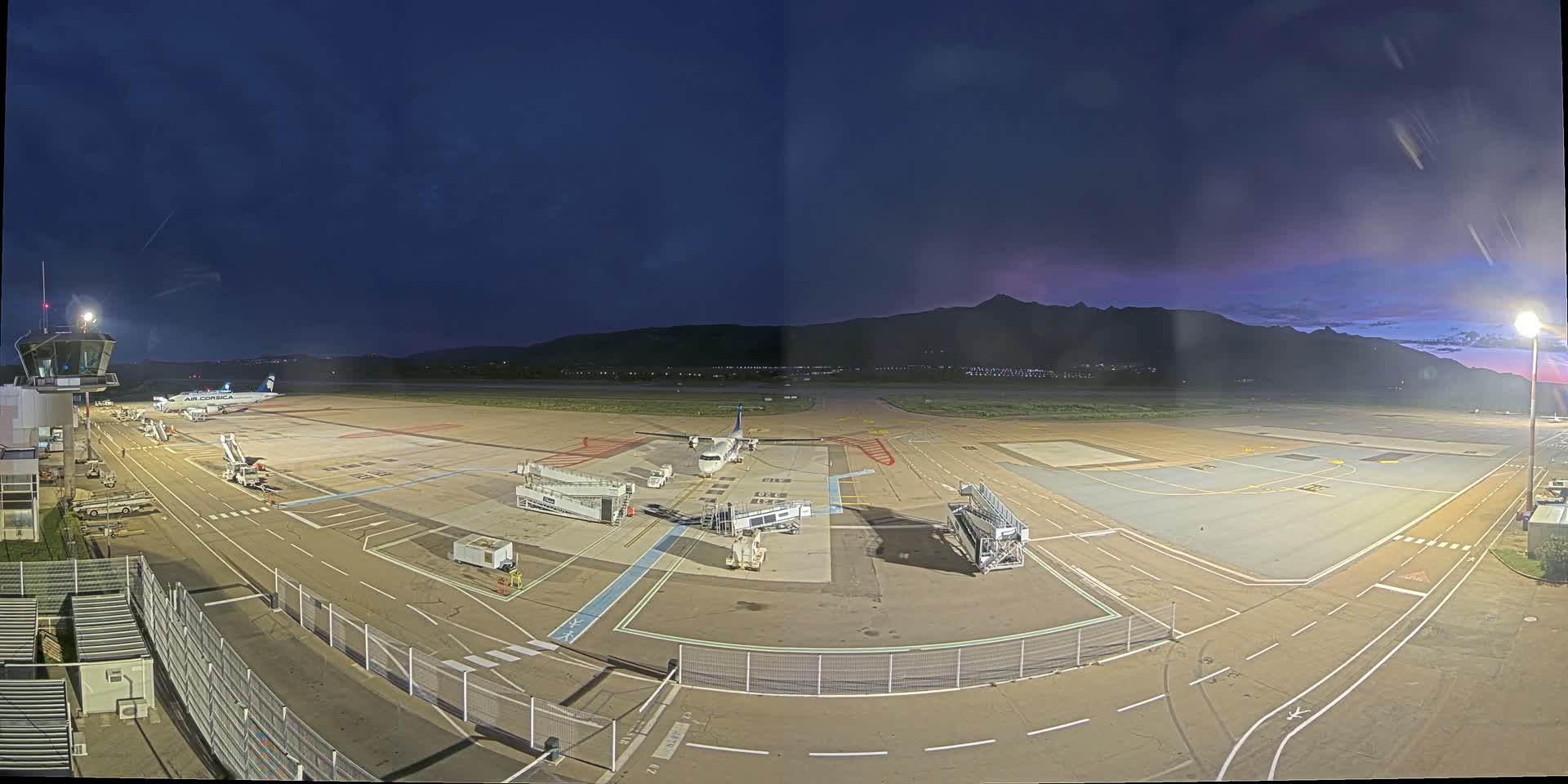 An airport tarmac at night under a twilight sky shows two airplanes parked at gates with jet bridges attached, along with support vehicles, a control tower, and distant mountains.