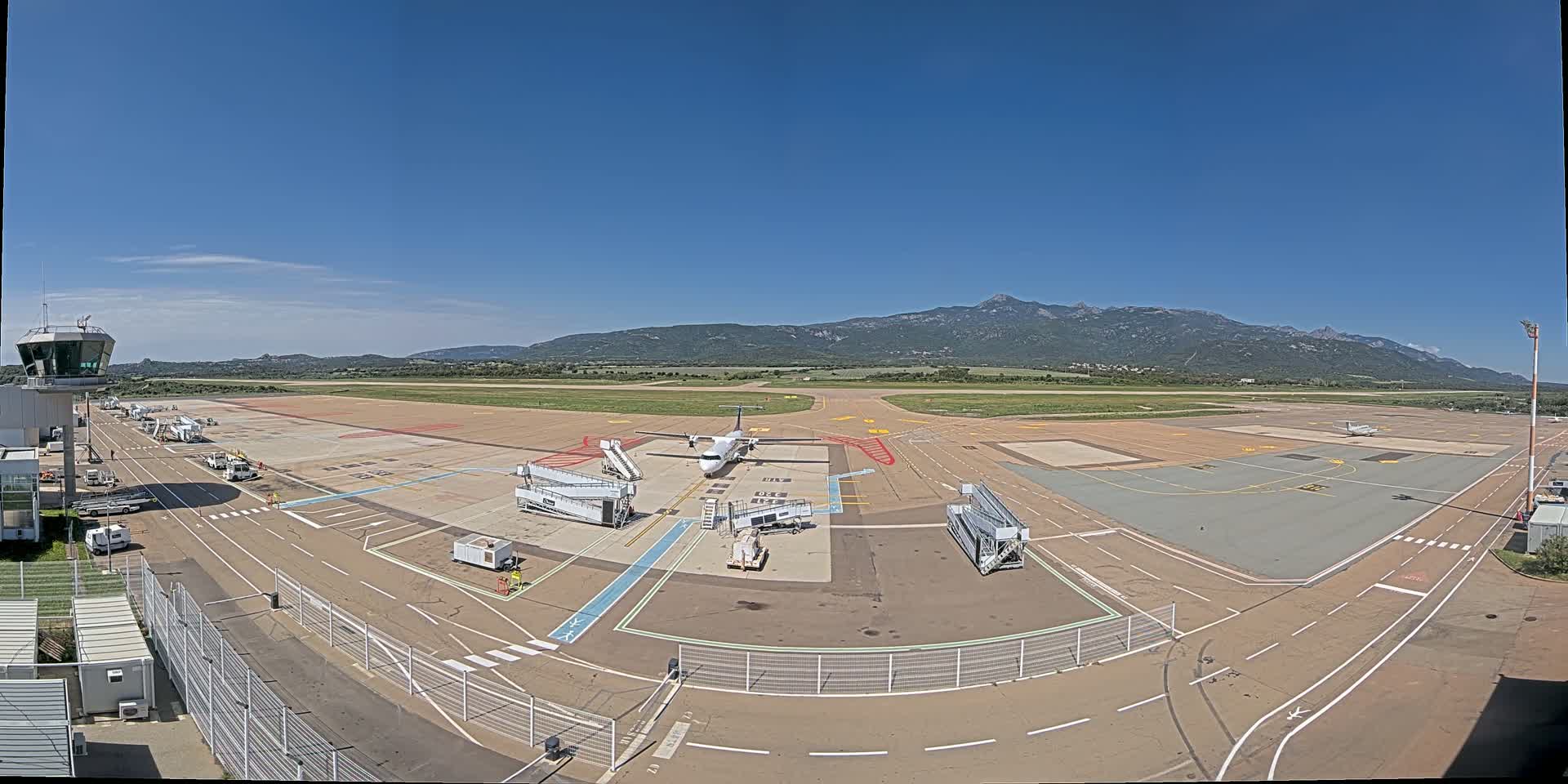 A small passenger airplane is parked at a gate of an airport tarmac on a clear, sunny day with mountains visible in the distance.