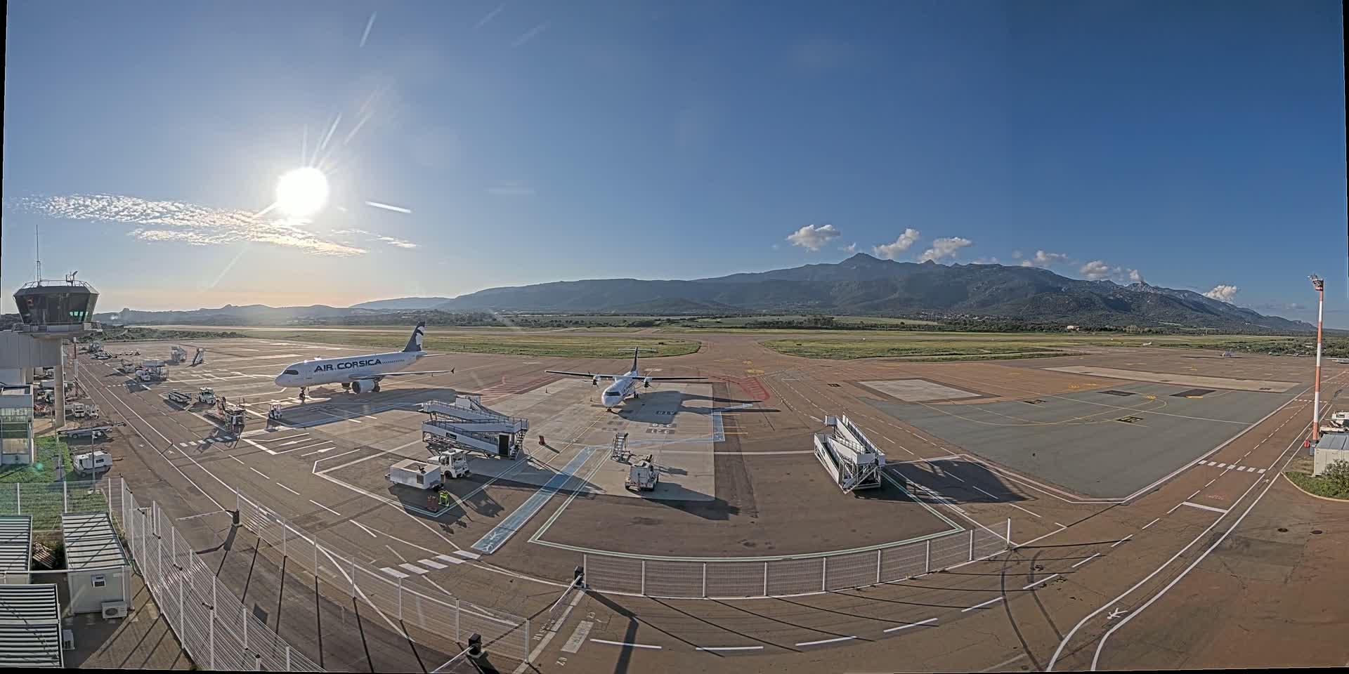 A sunny day at an airport shows two airplanes on the tarmac near gates, with mountains visible in the distance.