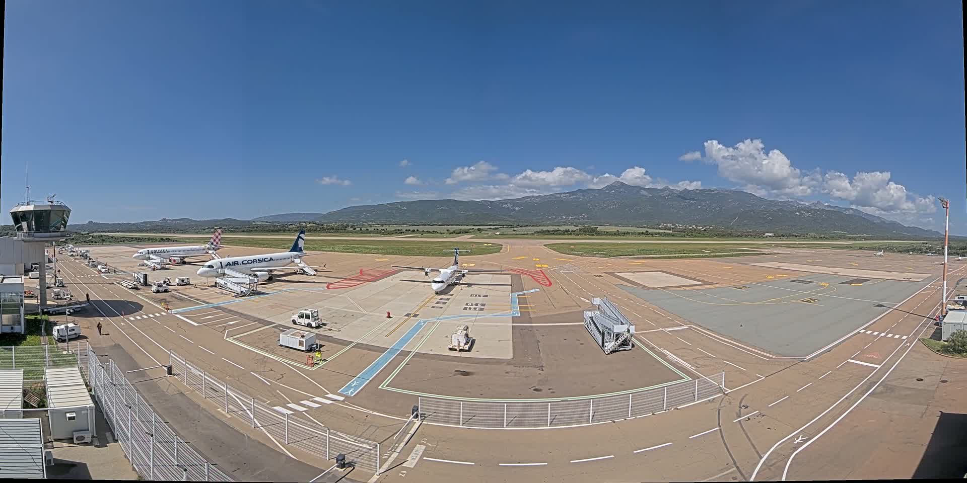 An airport tarmac with several airplanes parked near gates under a mostly sunny sky with some clouds and mountains visible in the distance.