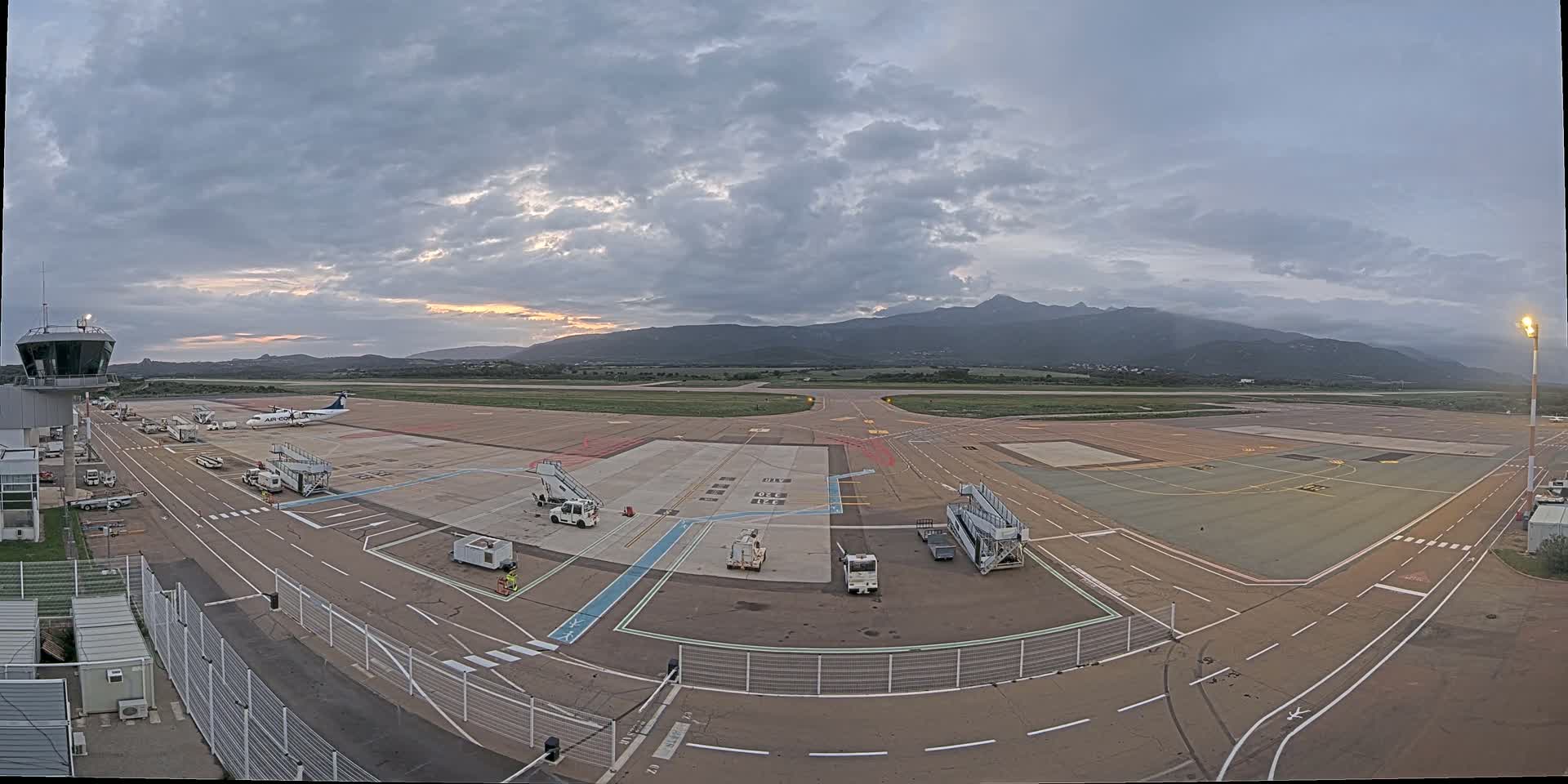 An airport tarmac with a plane and several ground support vehicles is visible under a cloudy sky, with mountains in the background.