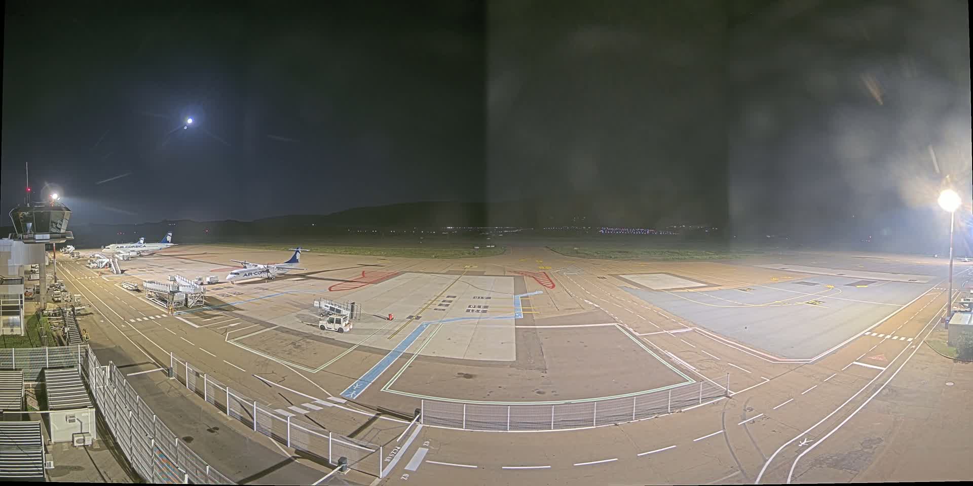A nighttime view of an airport tarmac with several airplanes parked, illuminated by bright lights under a moonlit sky.
