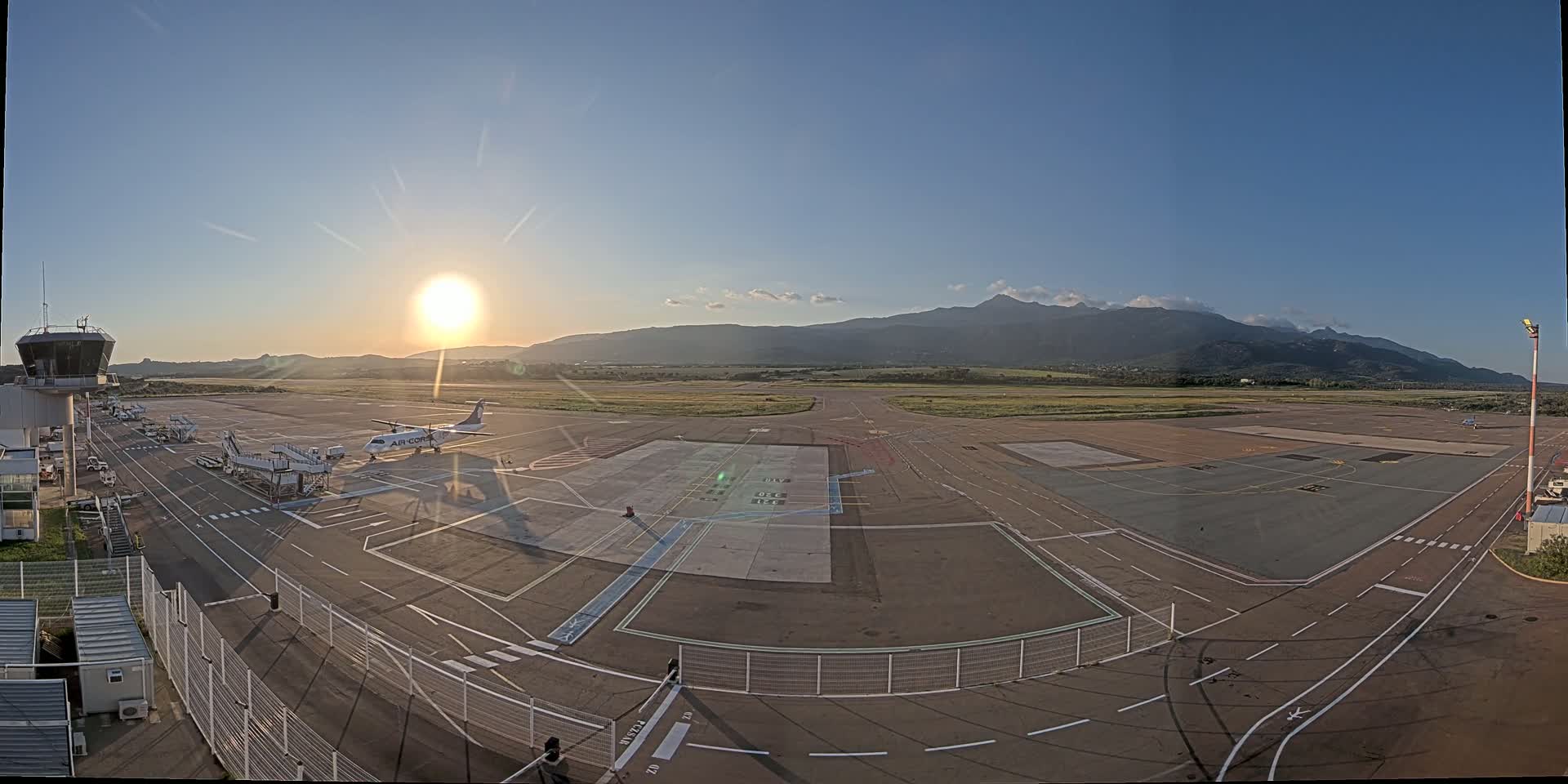 A mostly empty airport tarmac with a single airplane parked at a gate, under a bright, clear sunset sky with mountains in the distance.