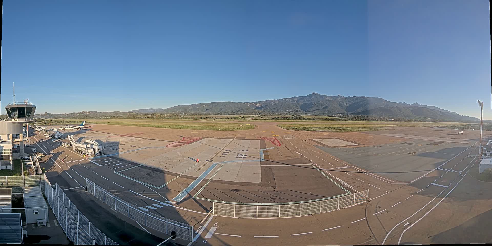 An airport tarmac with several airplanes, a control tower, and jet bridges is visible beneath a clear blue sky and in front of a mountain range.