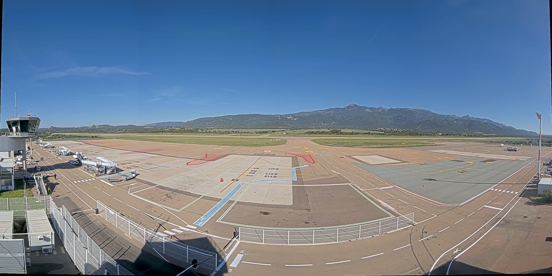 An airport tarmac with several jet bridges and a control tower, under a clear blue sky with mountains in the background.