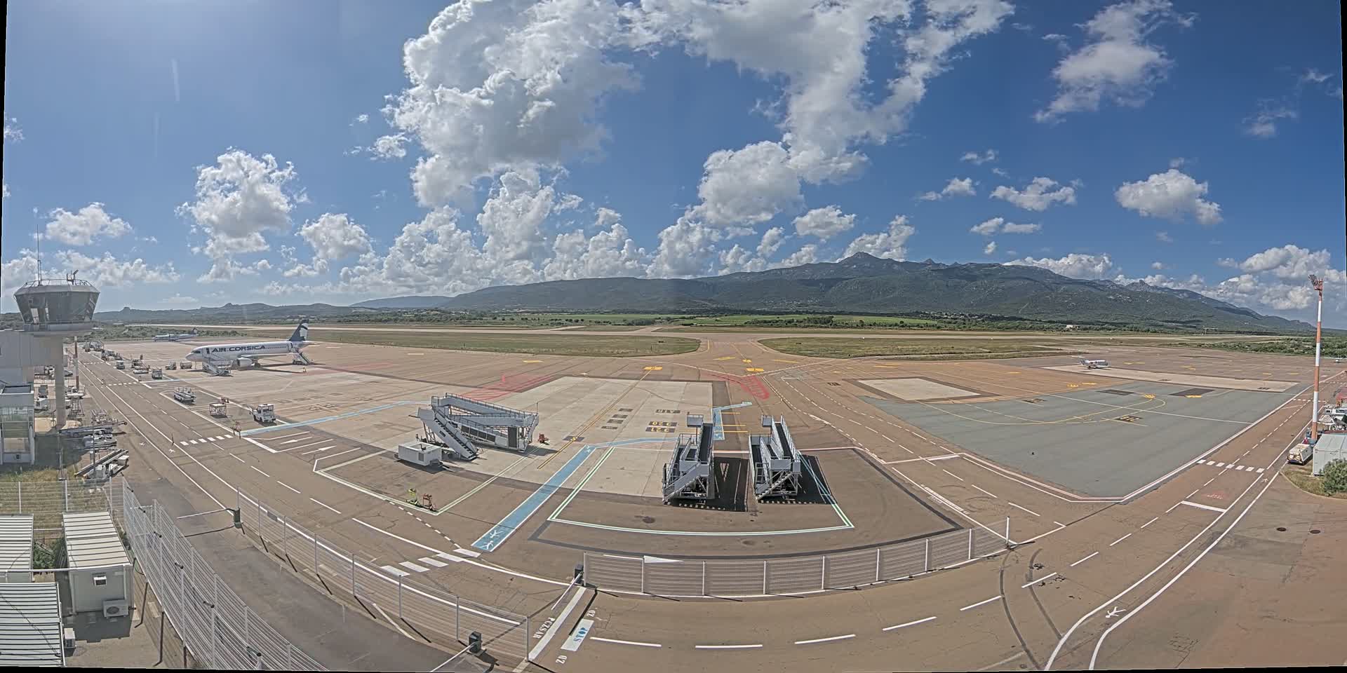 An airport tarmac with a single plane parked at a gate, several jet bridges, and a control tower, under a partly cloudy sky with mountains visible in the distance.