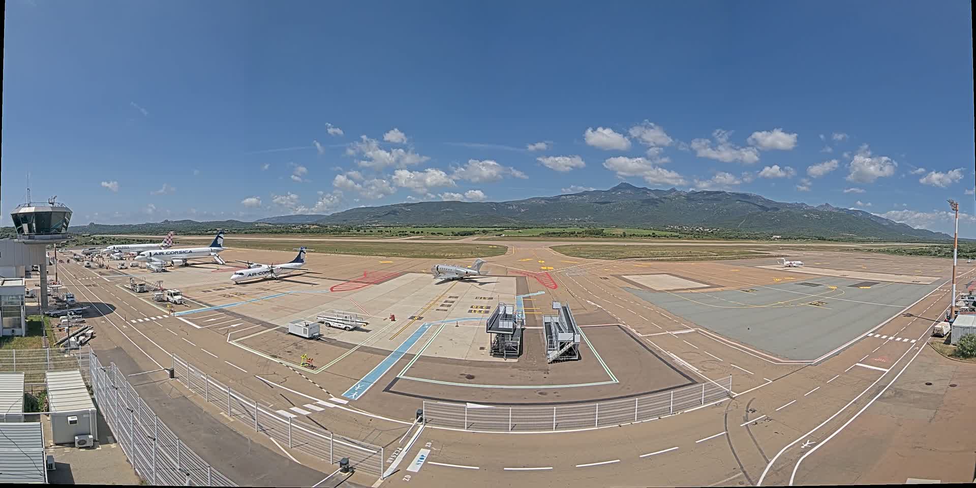 An airport tarmac with several airplanes parked, a control tower, and service vehicles under a mostly sunny sky with mountains in the background.