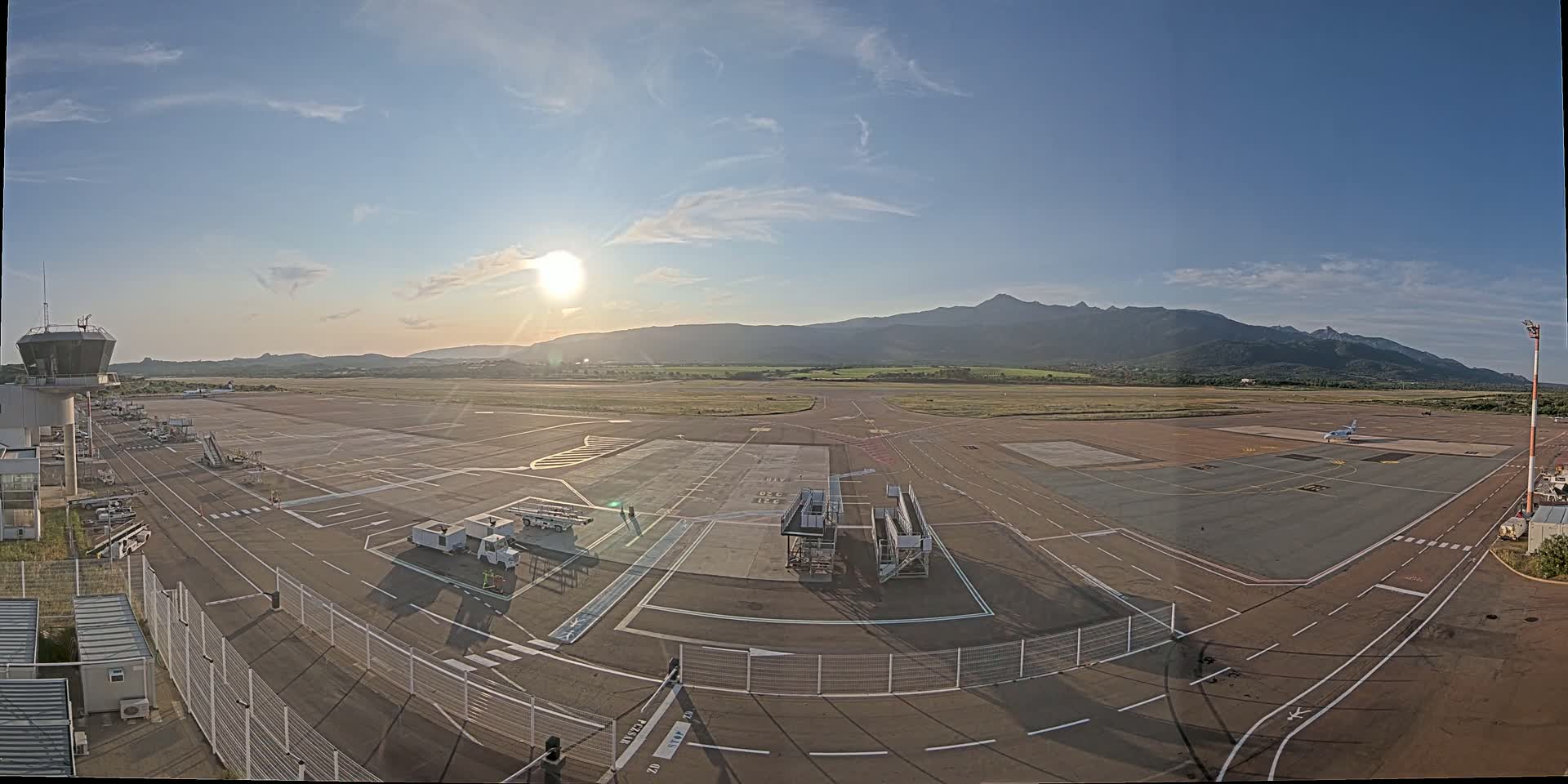 An airport tarmac with a control tower, several service vehicles, and a single airplane is visible under a mostly sunny sky, with mountains in the background.