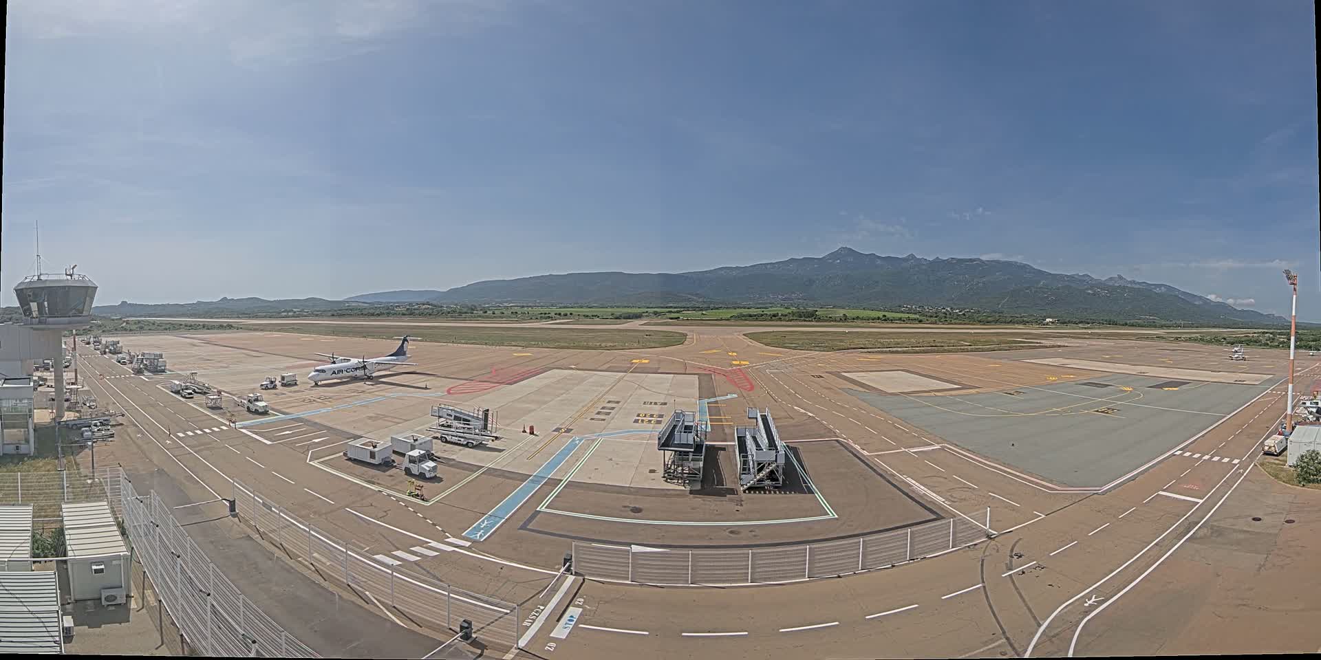 An airport tarmac on a sunny day with a single airplane parked near a terminal building and mountains visible in the distance.