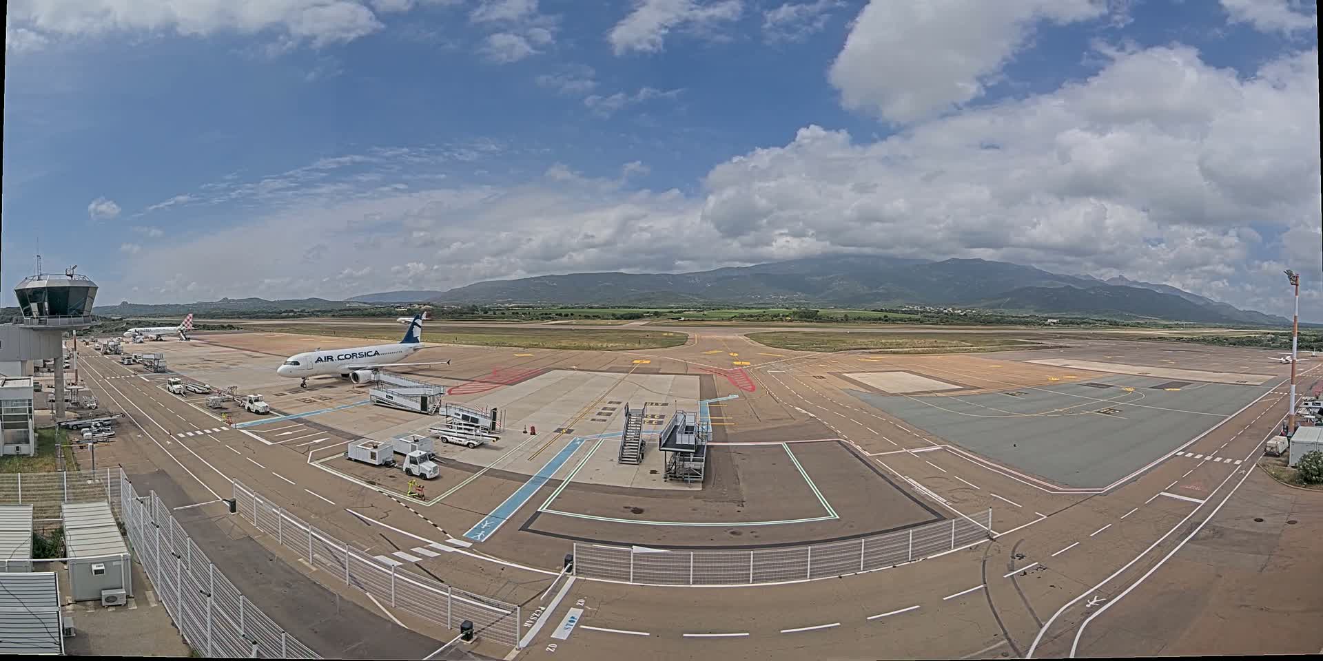 An airport tarmac with several airplanes, support vehicles, and a control tower under a partly cloudy sky, with mountains visible in the distance.
