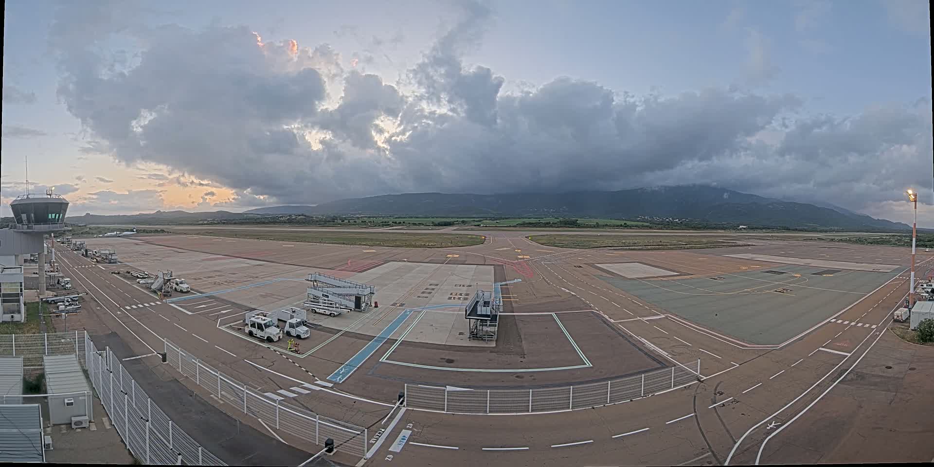 An airport tarmac with several service vehicles, a control tower, and a partly cloudy sky over distant mountains.