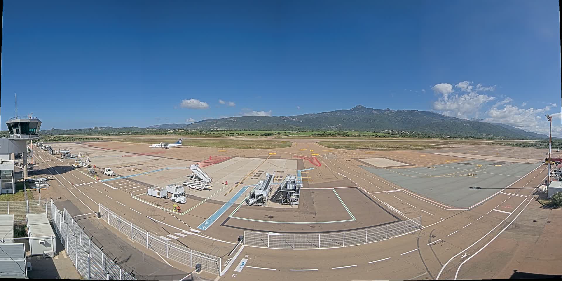 An airport tarmac with a single airplane, several ground support vehicles, and a control tower is visible on a sunny day with mountains in the background.