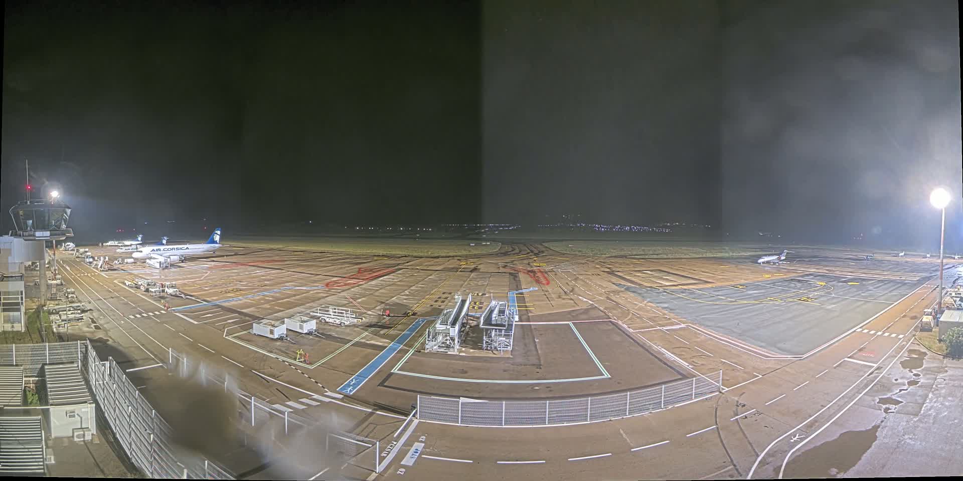 A nighttime view of an airport tarmac showing several airplanes parked, with wet pavement under a dark, hazy sky.