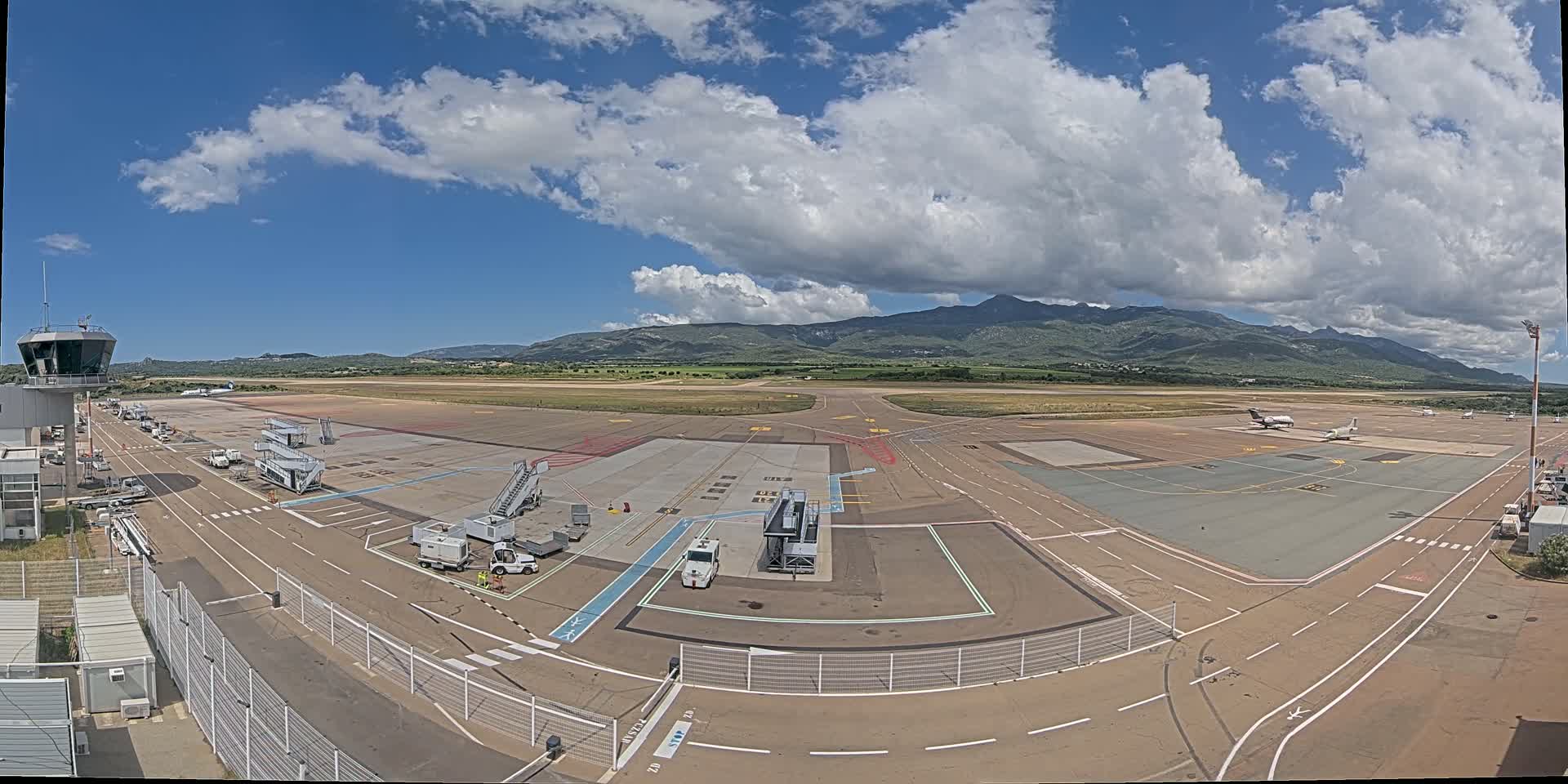 A mostly sunny day at an airport with several planes parked on the tarmac, jet bridges, support vehicles, and a control tower, with mountains visible in the background.