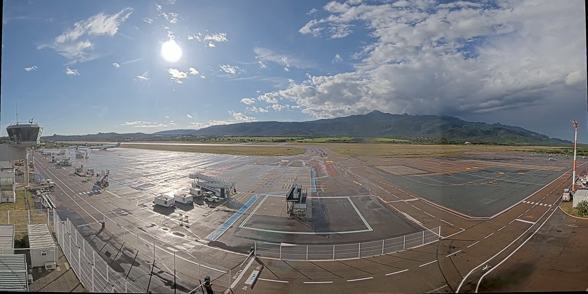 A wet airport tarmac with several planes, service vehicles, and a control tower, under a partly sunny sky with mountains in the distance.