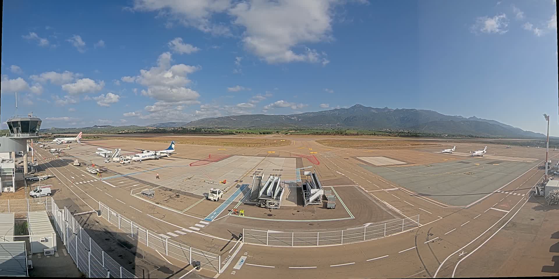 A mostly sunny day at an airport shows several planes parked on the tarmac with mountains visible in the distance.