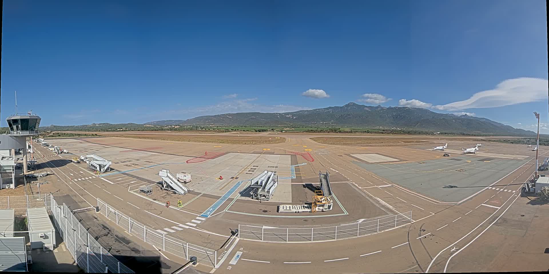 An airport tarmac with several planes and jet bridges, under a mostly clear blue sky with mountains in the distance.