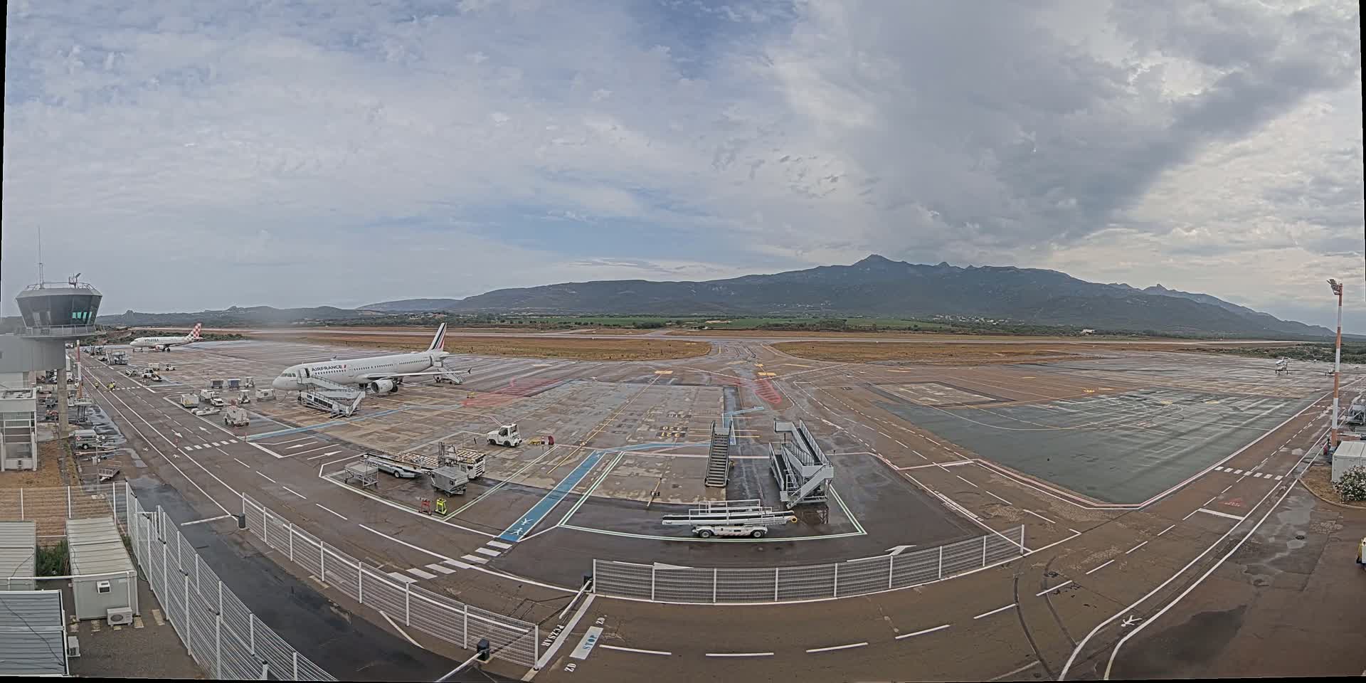A wet airport tarmac with several airplanes and support vehicles, under a cloudy sky with mountains in the background.