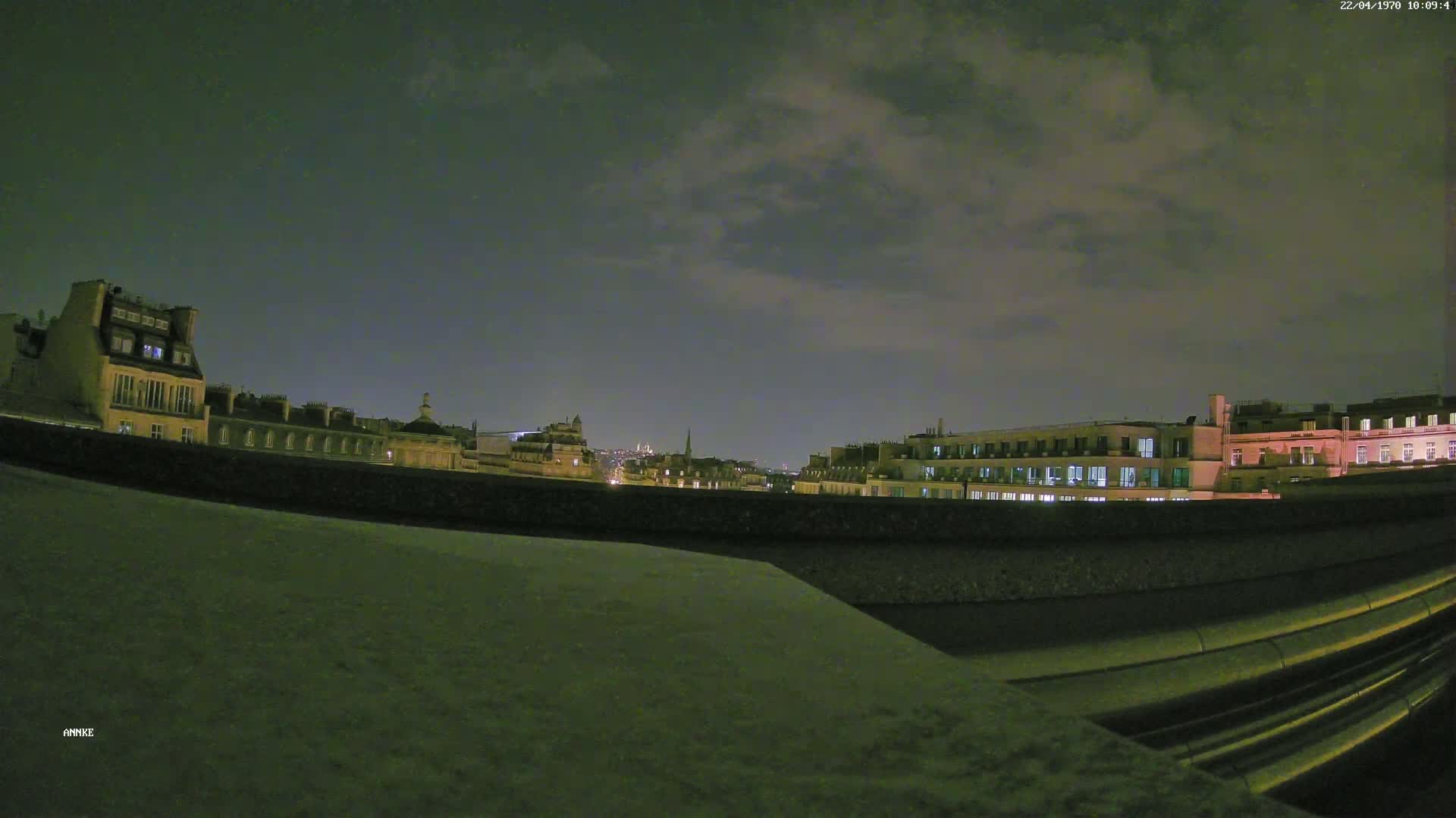 A nighttime view of city buildings under a cloudy sky, seen from behind a low wall.