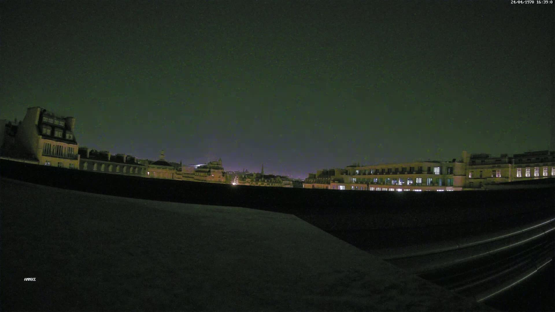 A nighttime view from a rooftop shows a cityscape of dimly lit buildings under a dark, clear sky.