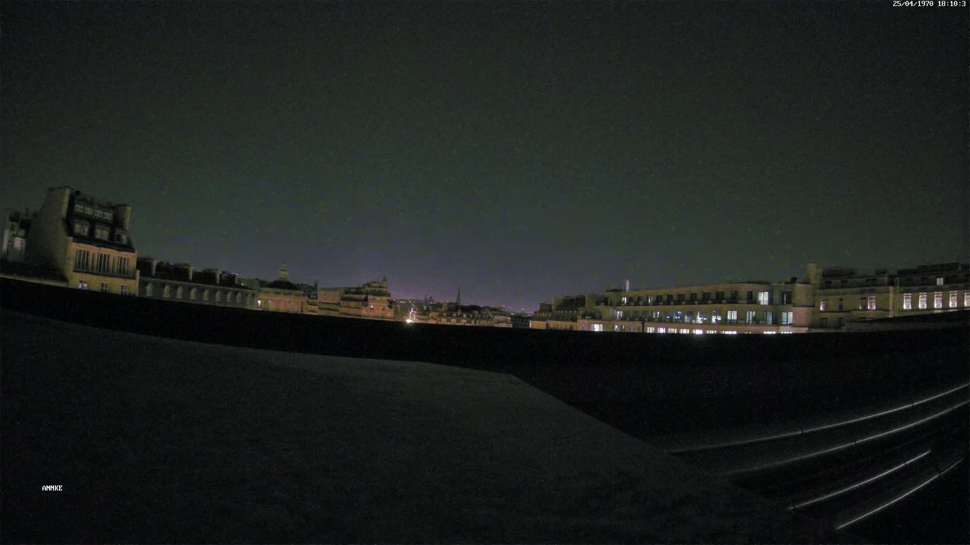 A nighttime view from a rooftop shows a cityscape of buildings with illuminated windows under a dark, clear sky.